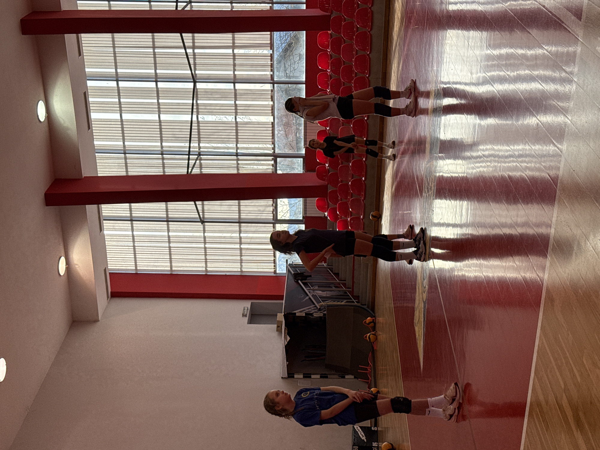 Several female athletes are performing warm-up exercises in an indoor sports hall. The hall features a wooden floor with red markings, red seating in the background, and a large windowed wall allowing natural light. The location is Etropole, Bulgaria. The athletes are wearing sportswear and knee pads, suggesting they are preparing for a volleyball game. Some volleyballs are on the floor.