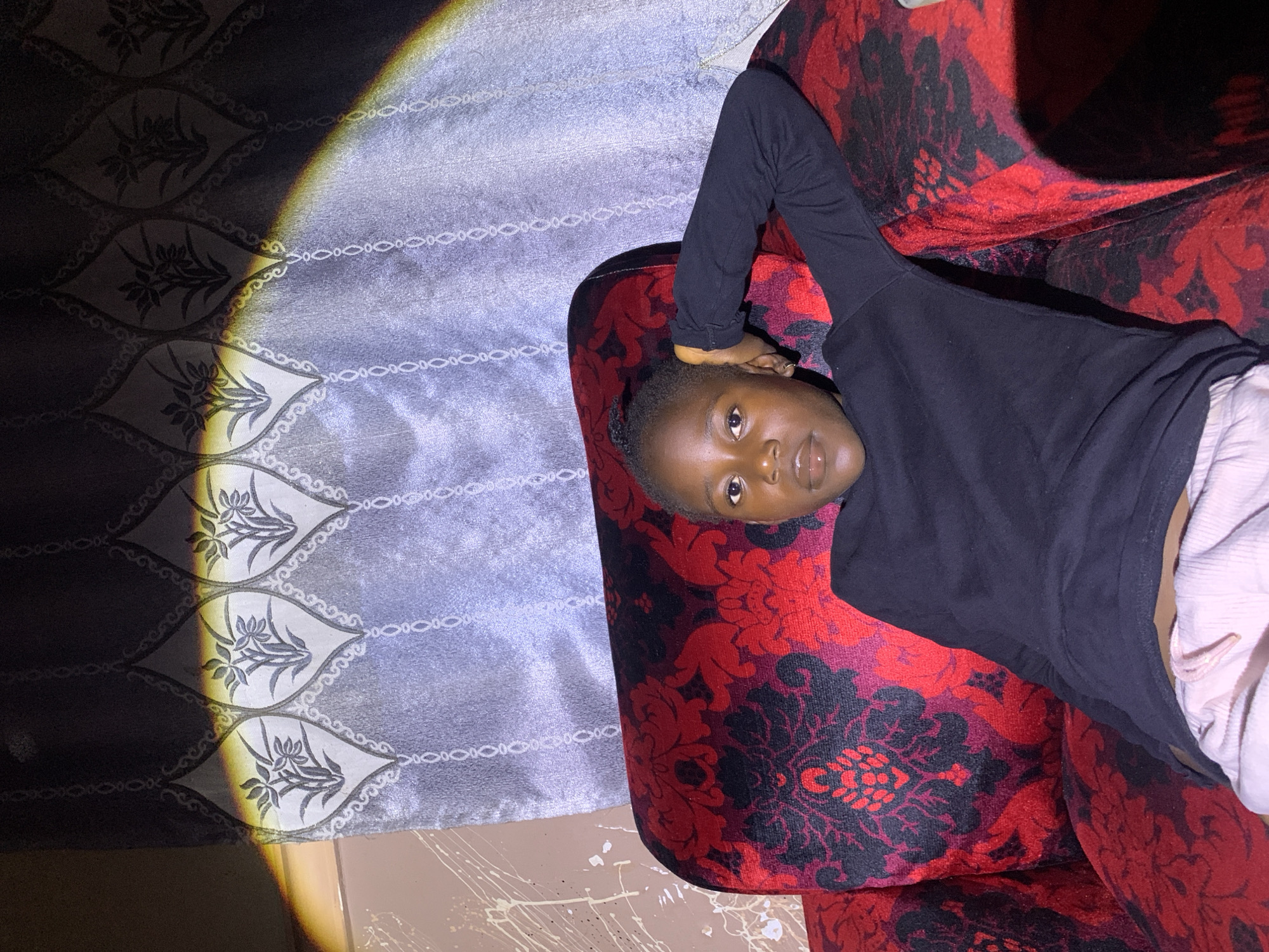 This is a media file captured in Russo, Nigeria. The scene features a young person lying on a red and black patterned sofa. The person is wearing a black long-sleeved shirt and light-colored bottoms. Their head is propped up on their arm, and they are looking towards the camera with a neutral expression.
To the left of the sofa is a grey curtain with a repeating pattern of stylized leaves and ornate borders. A bright light casts a distinct shadow and a yellowish-orange beam of light across the curtain, creating a dramatic contrast between light and shadow. The light suggests a time of day when a strong, directional light source, possibly natural sunlight or a powerful lamp, is present.
The floor beneath the sofa is visible and appears to be tiled, with some white paint splatters scattered across it. The overall impression is of an indoor setting, possibly a living room, with an artistic or dramatic lighting effect highlighting the textures and patterns of the environment and the subject. No visible text is present in the media file.