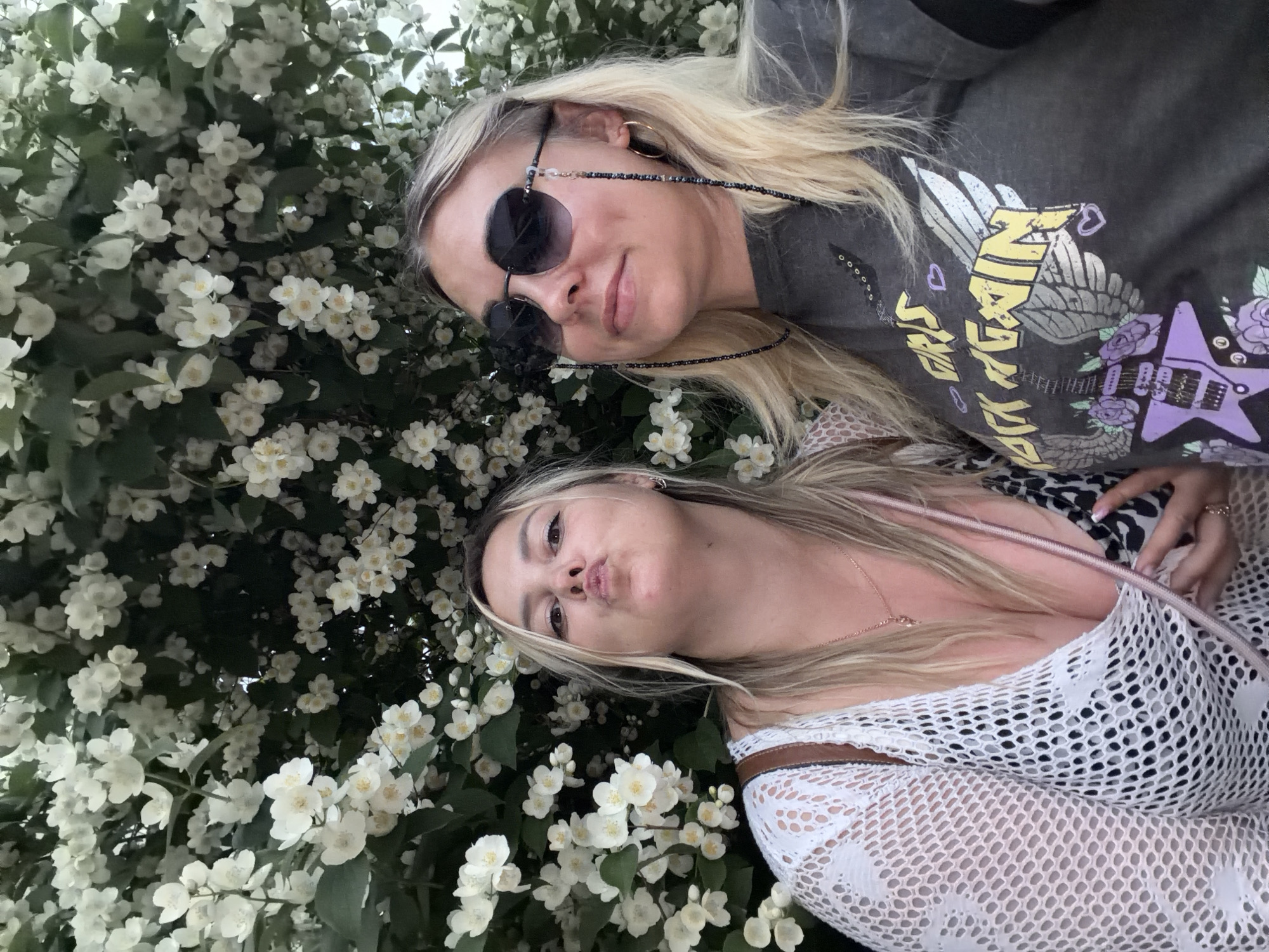 Two young women are posing for a selfie in front of a flowering bush. The woman on the left is puckering her lips and wearing a white mesh top over a black and white patterned bikini top. She has blonde hair and is wearing a necklace. The woman on the right is wearing sunglasses and a dark t-shirt with a graphic design. She has blonde hair and hoop earrings. The background is filled with white flowers and green leaves. The image is taken outdoors, likely in a garden or park.