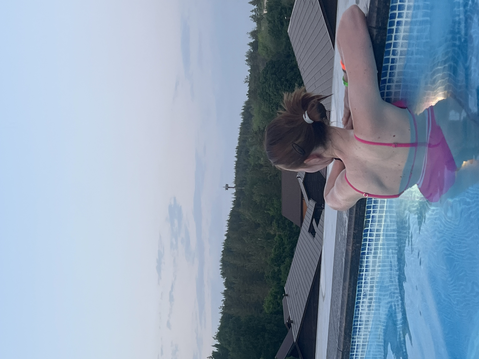 A woman is in a swimming pool. The pool's edge is tiled with blue and white mosaic tiles. The woman is wearing a pink swimsuit and has her hair tied back. Beyond the pool, there is a view of a forested area, and a building with a dark roof is visible to the right of the pool. A light-colored sky with clouds can be seen above the trees. There appears to be a tower structure rising above the forest in the background. There are no other readily identifiable objects in the image. No action or interaction beyond the woman in the pool is apparent. The location of the scene is not explicitly stated, but it is presumed to be in Khvalynsk, Russia.