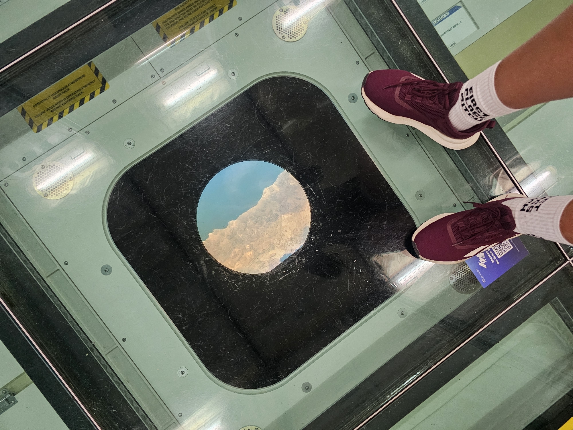 This image shows a first-person perspective of a person standing on a glass floor with a circular opening that reveals a view of rocky terrain and the sky below. The person is wearing burgundy athletic shoes with white soles and white socks with black text. The glass floor is part of a structure with a teal-colored frame. Yellow warning labels with black stripes are visible on the frame, and there are ventilation grates. A blue QR code sticker with the text "SPACE VIEW" is also visible on the glass floor. The location appears to be an observation deck or a similar attraction. The provided information indicates this is the city of Mukilteo, United States.