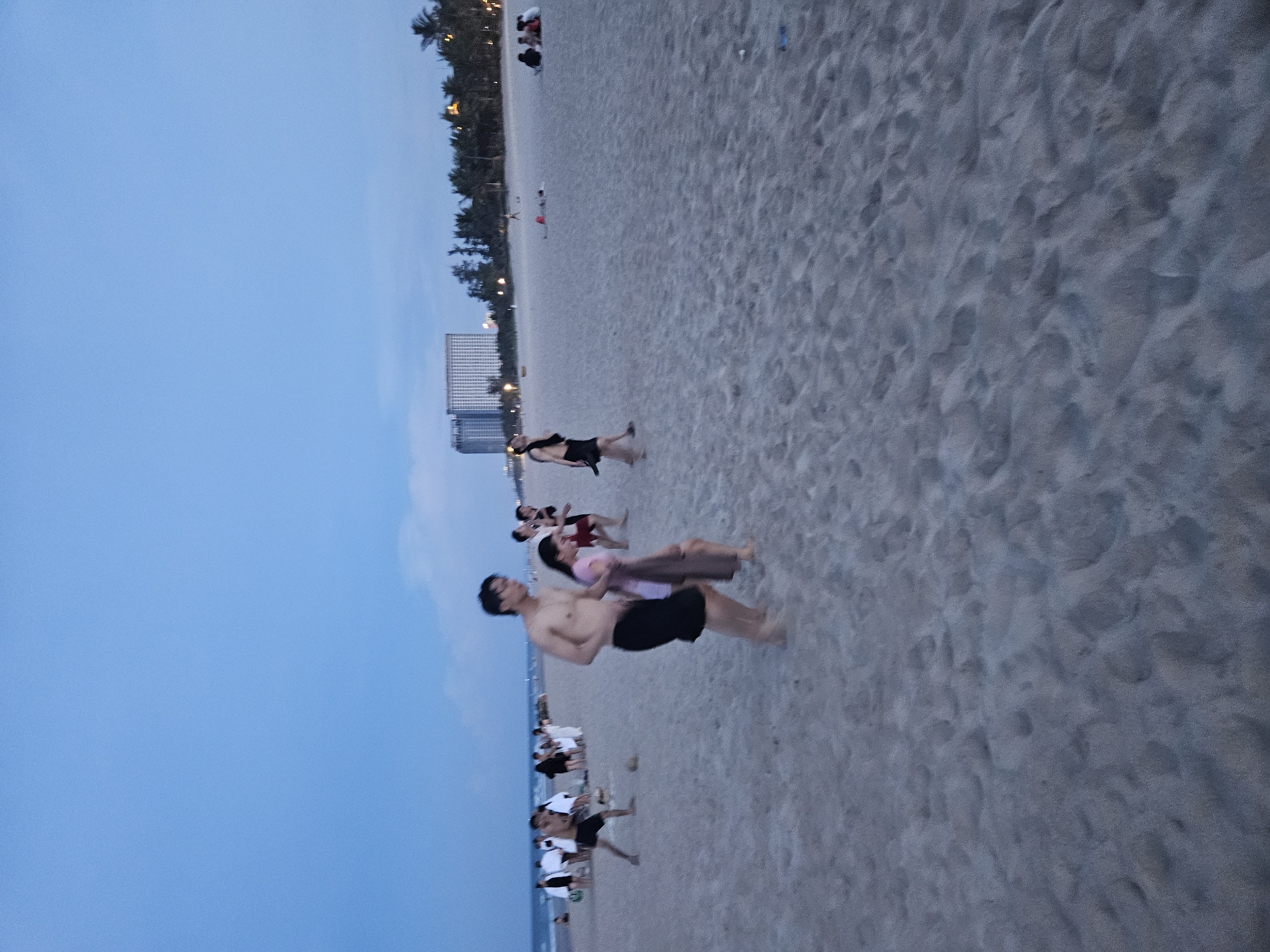 At dusk on a sandy beach in Đà Nẵng, Vietnam, several individuals are present. In the foreground, a shirtless man wearing dark shorts walks with a woman in a pink top and brown wrap. Behind them, other beachgoers are scattered across the sand. Some are standing in groups, while others are seated or lying down. In the background, tall buildings and palm trees line the coast, illuminated by lights. The sky is a deep blue, indicating the transition from day to night. The overall atmosphere is that of a relaxed evening at a popular tourist destination.