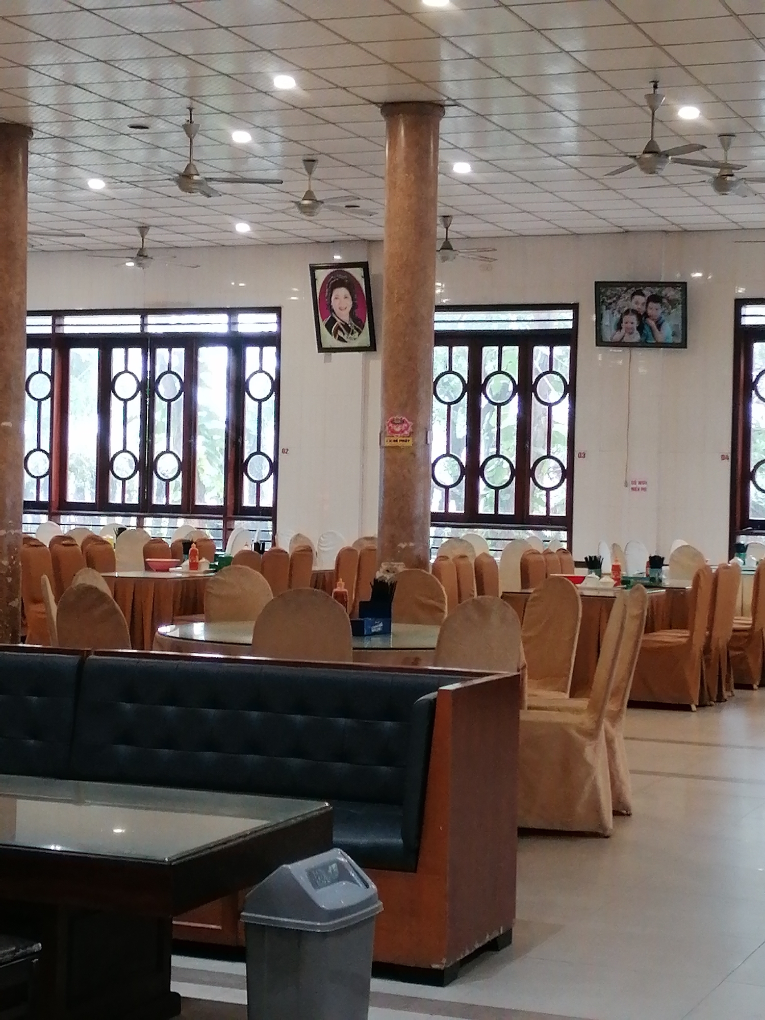 The image shows the interior of what appears to be a dining hall or restaurant in Chu Văn An Ward, Vietnam. The space is large, with numerous round tables covered in cloths. Chairs, also draped in fabric, surround each table. The seating area extends into the distance, indicating a substantial capacity. The furniture has an older, traditional style.
Visible are large windows with decorative circular patterns, allowing natural light into the room. Two framed pictures hang on the walls, one showing a person, and the other showing a family. Above, the ceiling features a grid pattern with multiple ceiling fans.
In the foreground is a dark-colored booth or sofa. A glass-topped table sits in front of it. Next to the table, there is a trash can. The overall impression is that of a functional, moderately decorated space, likely used for large gatherings or events.