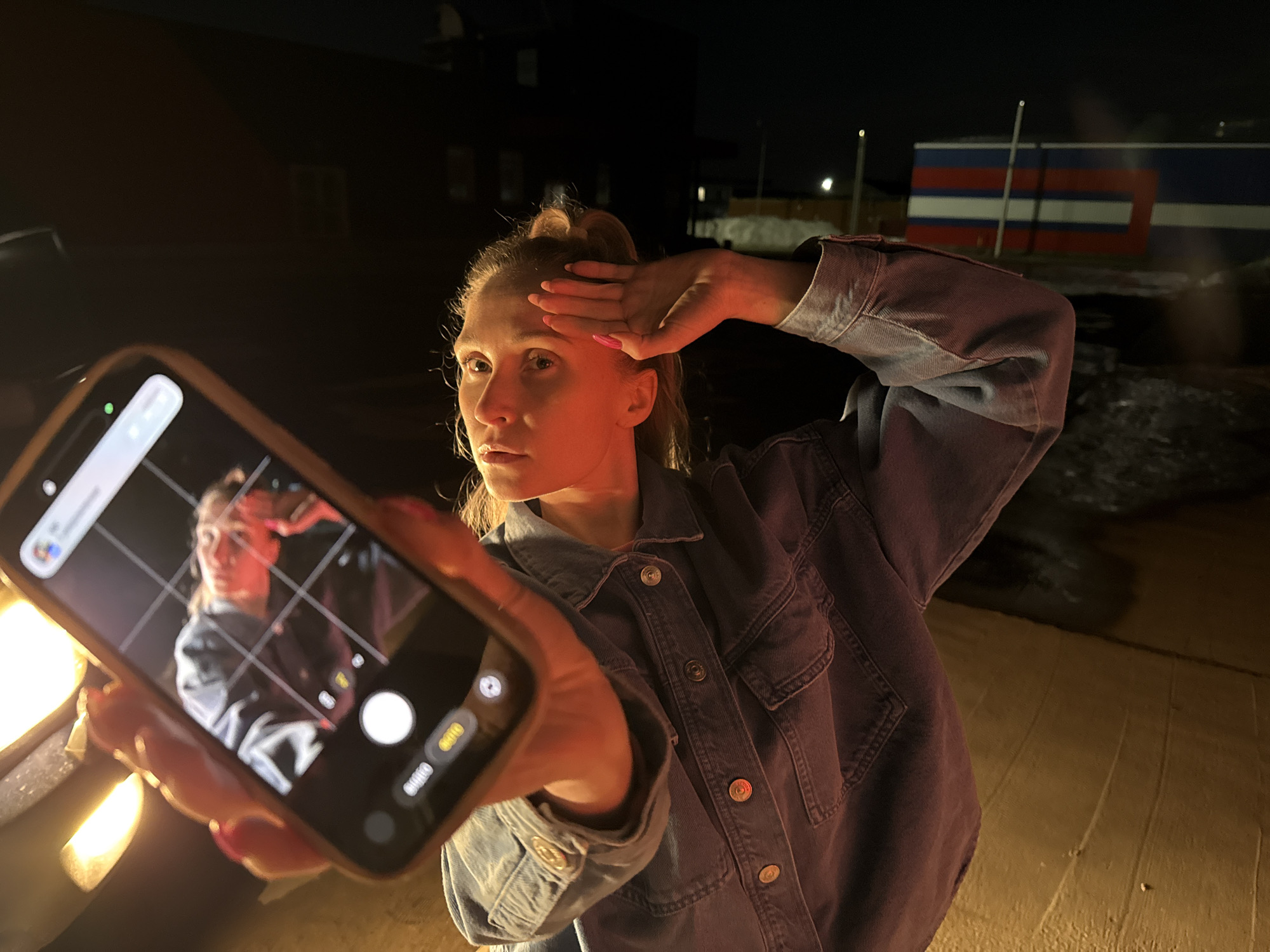A woman with fair skin and blonde hair pulled into a high bun is posing for a selfie at night. She is wearing a blue denim jacket and holding a smartphone in her left hand, with the screen displaying a reflection of herself mimicking her pose. Her right hand is raised to her forehead.
The background is dark, with a building displaying red, white, and blue horizontal stripes (suggesting the Russian flag) visible in the distance. There are also a few street lights visible. The scene is illuminated by a light source to the left of the smartphone, which causes the warm glow on the woman's face and hands.
Based on the presence of a building displaying red, white, and blue horizontal stripes, this image was taken in Chaikovsky, Russia.