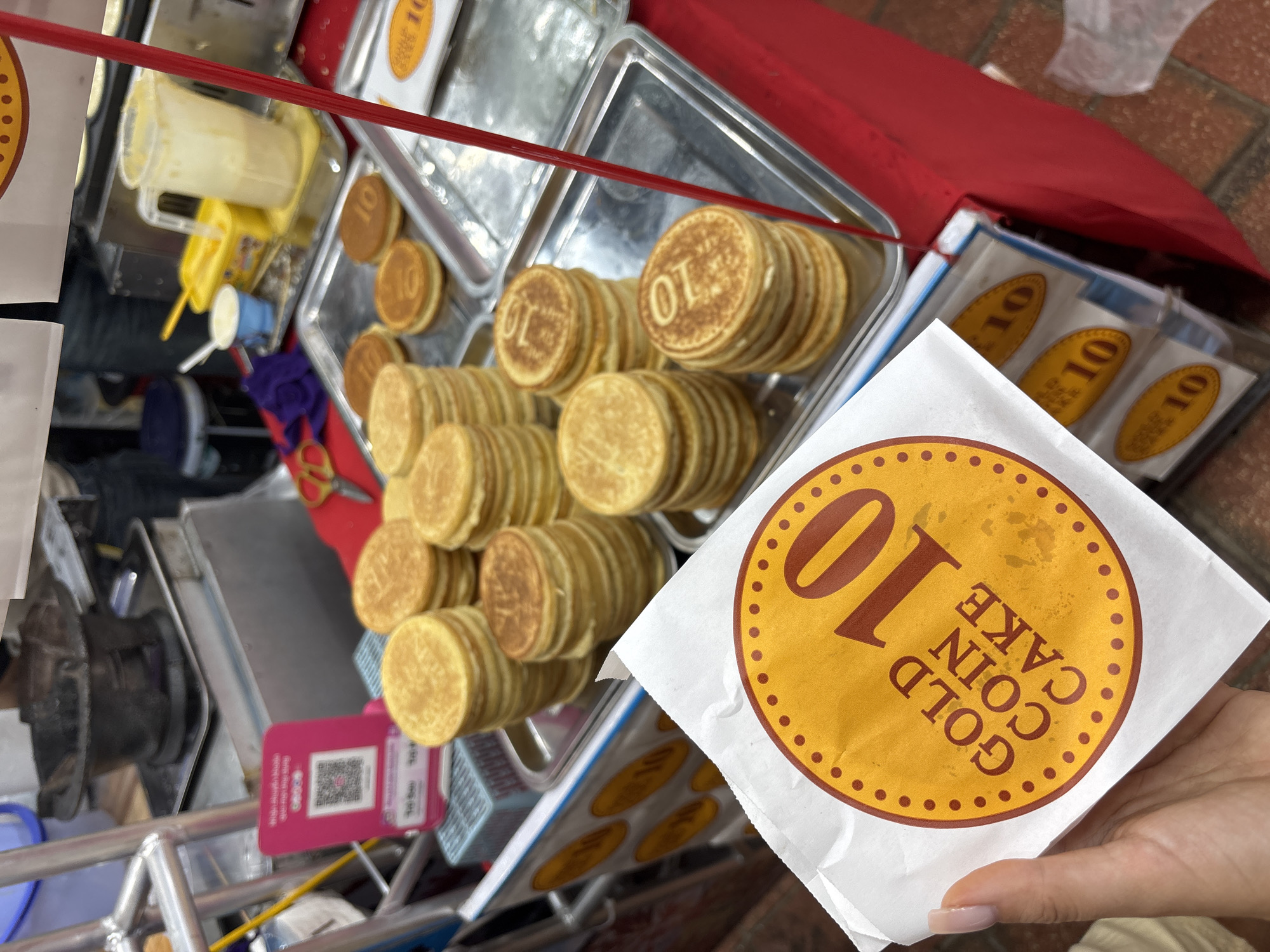 This image captures a vibrant street food stall, likely in Đà Nẵng, Vietnam, showcasing freshly made "Gold Coin Cakes." Numerous small, golden-brown, coin-shaped cakes are neatly stacked in rows on two rectangular metal trays, visible in the center and right of the frame. Each cake is embossed with the number "10".
In the foreground, a person's hand, with neatly manicured nails, holds a white paper bag. This bag serves as packaging for the cakes and features a prominent circular golden graphic with a dotted border. Inside the circle, the text "GOLD COIN CAKE" is written vertically, accompanied by a large, bold "10". Several other identical paper bags are folded and ready for use on a lower shelf of the stall, slightly out of focus.
The street food vendor's setup includes a red awning or cover along the top and right side of the image. Behind the display of cakes, various vendor supplies are visible: a clear plastic pitcher containing a pale yellow liquid (possibly batter or a topping), a yellow squeeze bottle, and other cups with spoons, suggesting tools for food preparation. On the lower left side of the stall, a pink sign with a QR code is attached, indicating modern digital payment options. The ground below the stall is tiled.
The scene portrays a typical daytime street food operation, with the cakes prepared and ready for sale. The hand holding the packaged item suggests an active transaction, either a customer receiving their order or a vendor preparing it. The number "10" on the cakes and packaging could represent a price unit, a specific flavor, or a branding element. The overall atmosphere is that of a bustling, informal market or street food environment.