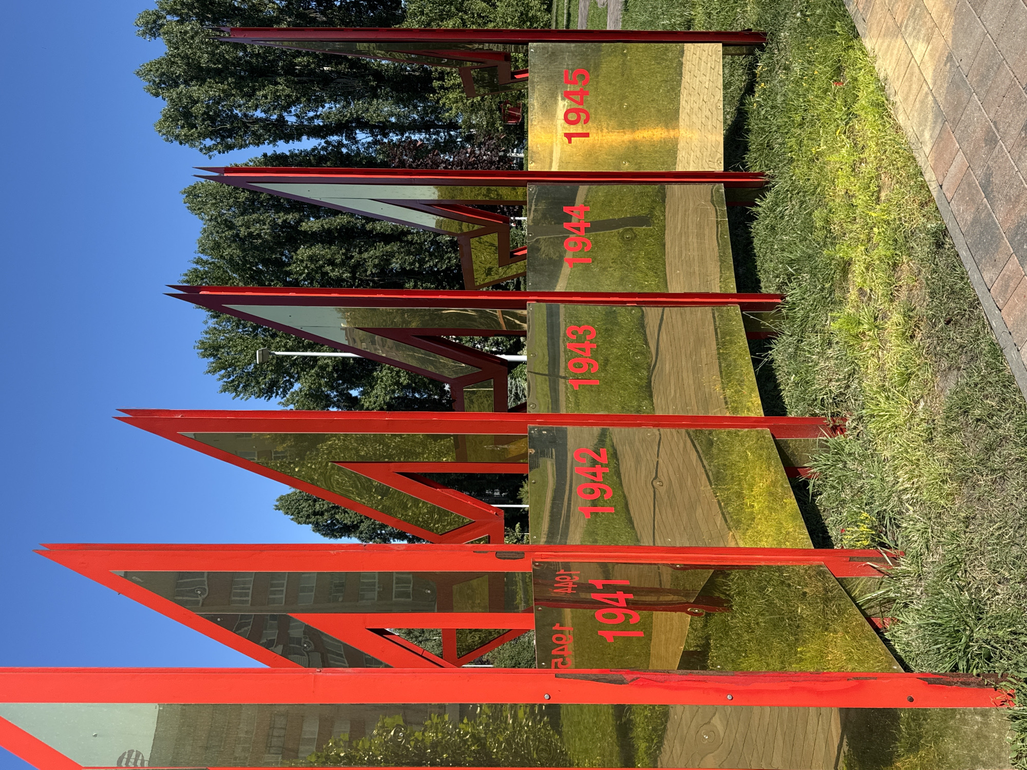 This image shows a memorial structure made of red metal frames and golden reflective panels, located in Nizhny Novgorod, Russia. The memorial is in the shape of stylized letters, possibly forming a word related to a historical event or anniversary. The reflective panels display the reflections of trees, a building, and the blue sky. Inscribed on some of the panels are the years "1941", "1942", "1943", "1944", and "1945" in red lettering, indicating a commemoration of World War II or a related period. The memorial is situated outdoors, in a grassy area with a paved walkway in the foreground. There are no people visible in the image.