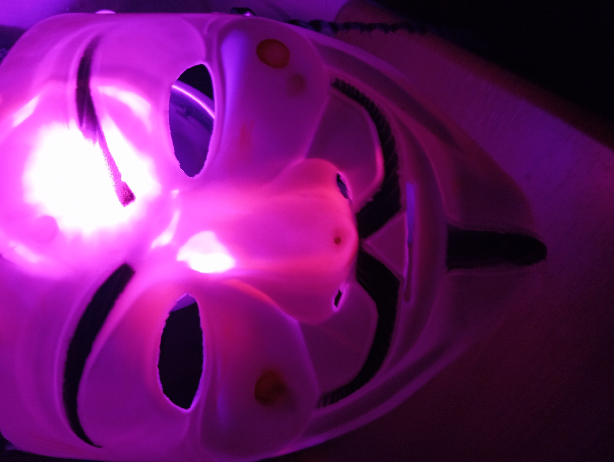 The image is a close-up, low-angle shot of a white Guy Fawkes mask illuminated by a bright purple light from behind, creating a silhouette effect and highlighting the mask's features. The mask has dark openings for the eyes and mouth, and circular orange markings on the cheeks. The lighting is intense and uneven, with the brightest spot directly above the mask. The background is dark and out of focus, with hints of purple light reflecting on surfaces. There is no visible indication of the city of Zaporizhzhia, Ukraine, or any specific location.