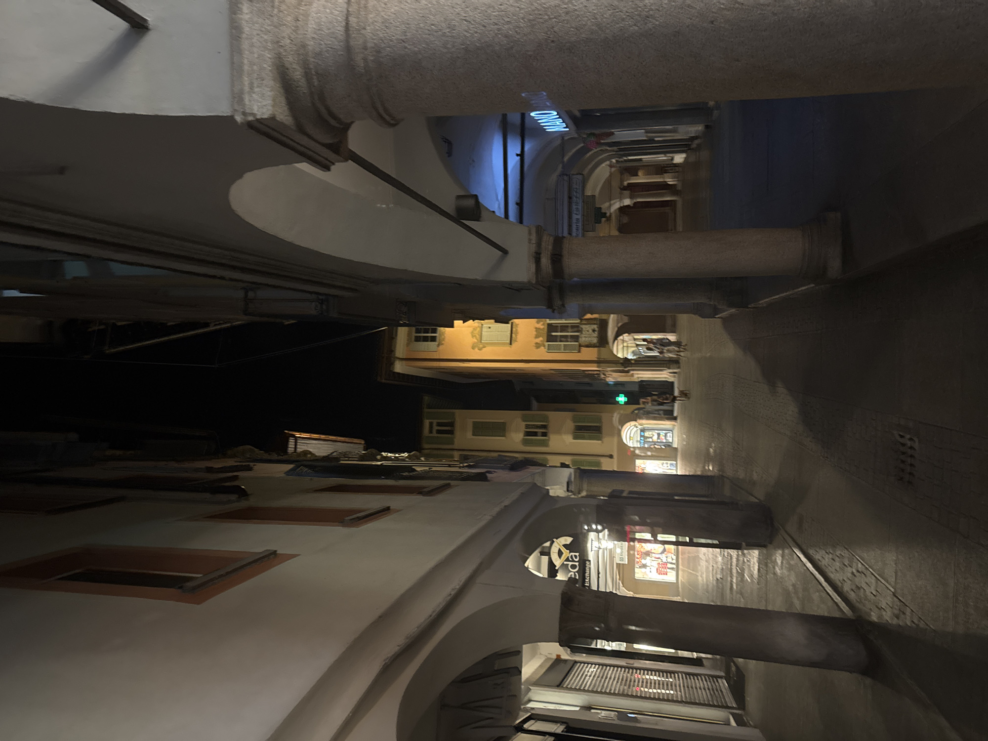 The image depicts a street scene at night in Lugano, Switzerland, characterized by an arcade with stone columns and arches on the right side, and buildings with windows and storefronts on the left. The lighting is dim, with artificial lights illuminating the shops and casting shadows. Several shop signs are visible, including "eda" and "MANO" in neon. The street surface is paved with cobblestones. Two individuals are walking in the distance on the left side of the street. The architecture suggests an older European city.