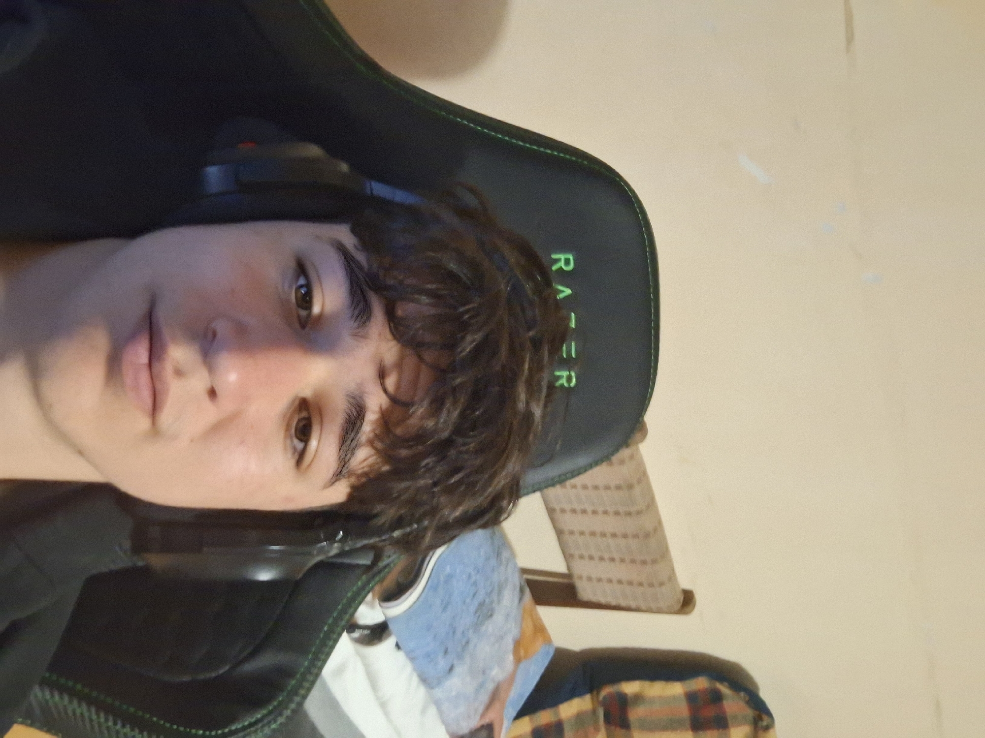 A person with brown hair is sitting in a black and green Razer gaming chair. The person is wearing headphones and facing the camera. The background is a beige wall and part of what appears to be a bed with a blue and white blanket, and a chair with a patterned covering. It's not possible to determine the location in Burgas, Bulgaria from this image alone.