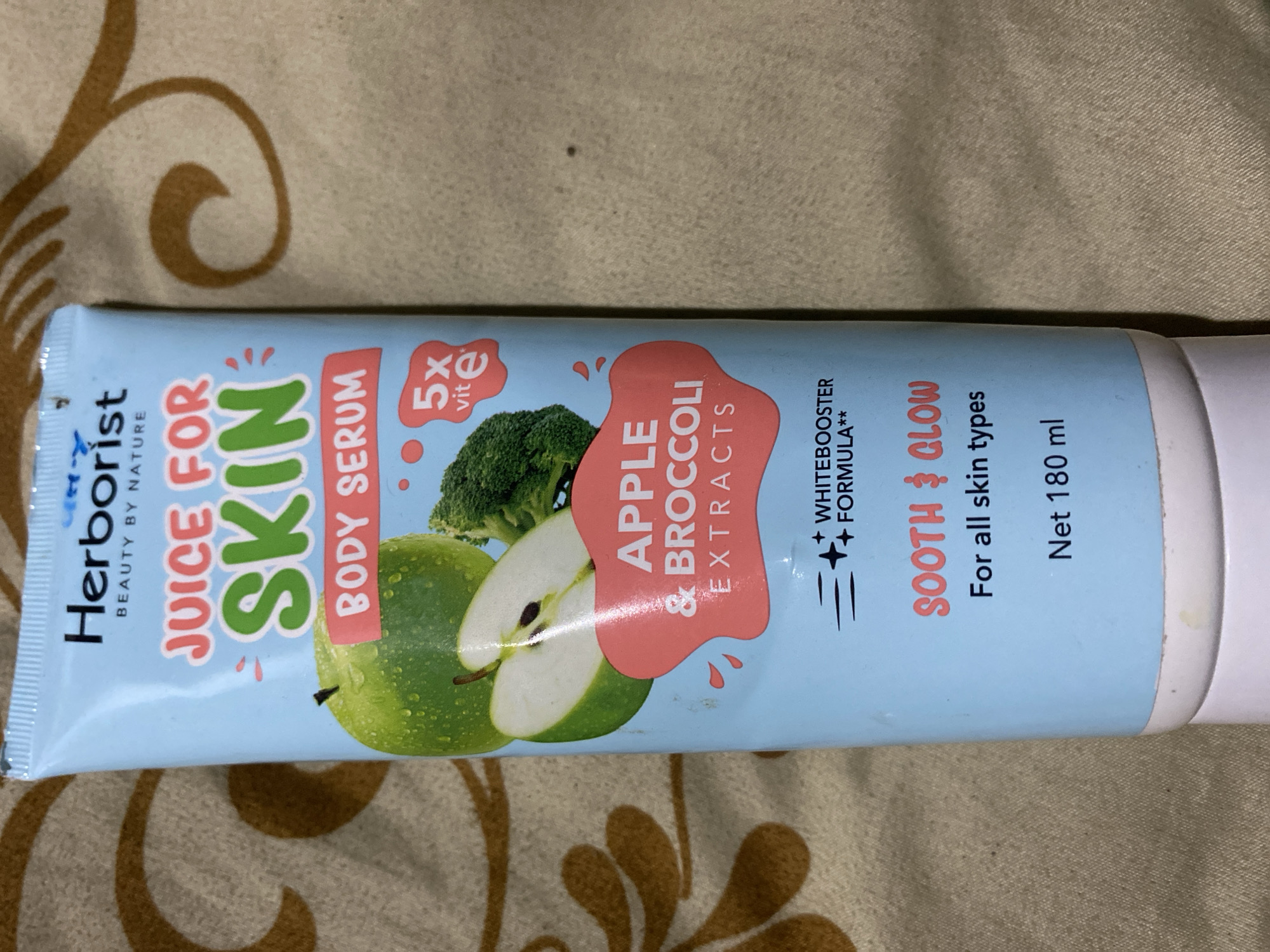 A light blue tube of Herborist Juice for Skin Body Serum with Apple & Broccoli Extracts is laid on a brown textured fabric. The tube is mostly upright, with its cap at the bottom. The label prominently features "Herborist," "BEAUTY BY NATURE," "JUICE FOR SKIN," and "BODY SERUM." An illustration of an apple and broccoli is on the front, along with a "5x vit e" callout. Below this, it says "APPLE & BROCCOLI EXTRACTS." Further down, there's "WHITEBOOSTER FORMULA**" and "SOOTH & GLOW For all skin types." The net content is "Net 180 ml." The background is a brown fabric with a subtle repeating pattern. The image was taken in Ta', Indonesia.