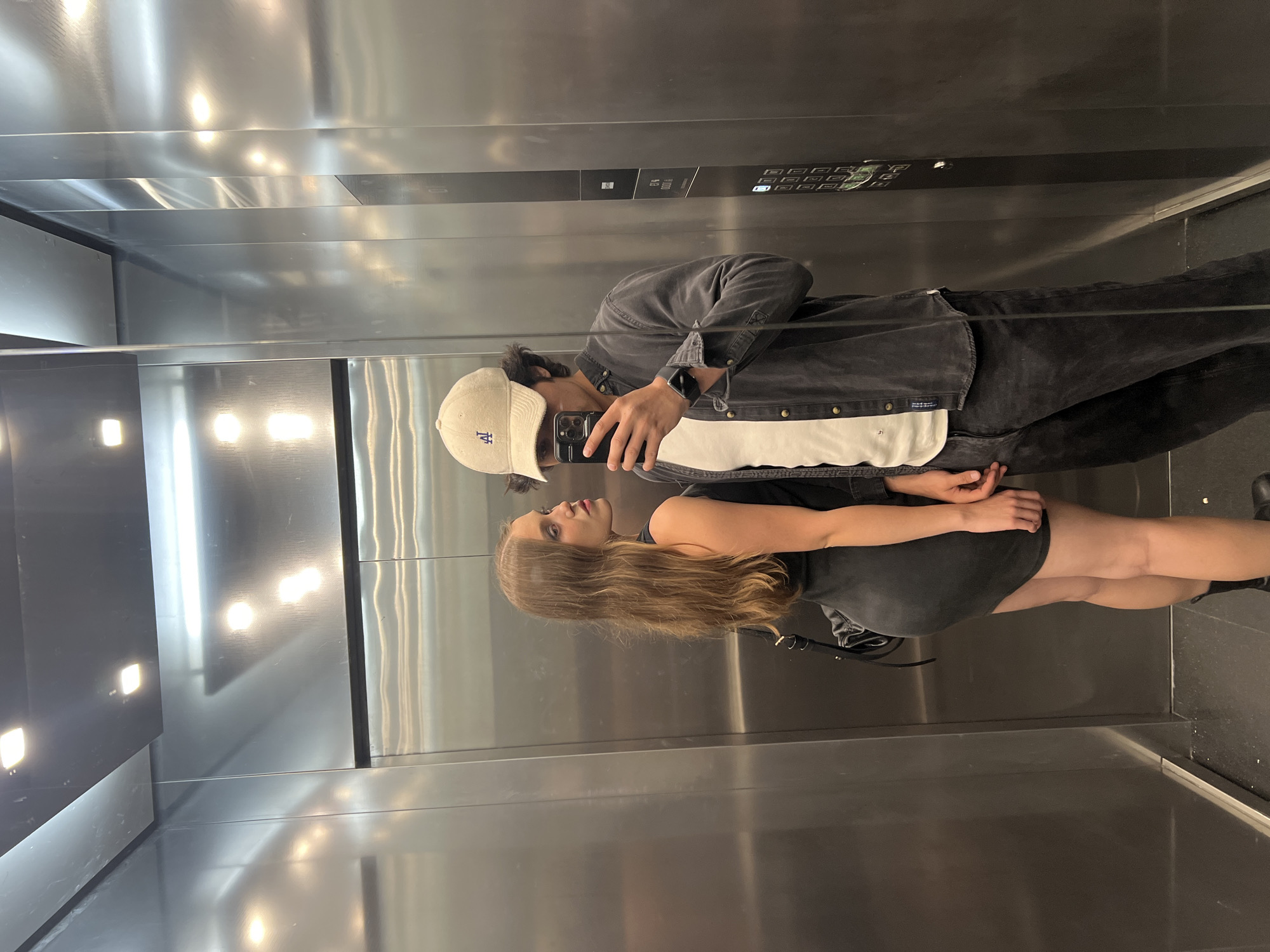 A man and a woman are standing in an elevator with shiny metal walls, taking a mirror selfie. The man is wearing a white baseball cap with the LA logo, a grey button-up shirt over a white t-shirt, and dark jeans. He is holding a cell phone up to the mirror to take the picture, with his watch visible on his left wrist. The woman is standing slightly in front of him, wearing a dark dress and boots. The elevator control panel is visible above and behind them. The location is likely in Mexico City, Mexico, based on the metadata.