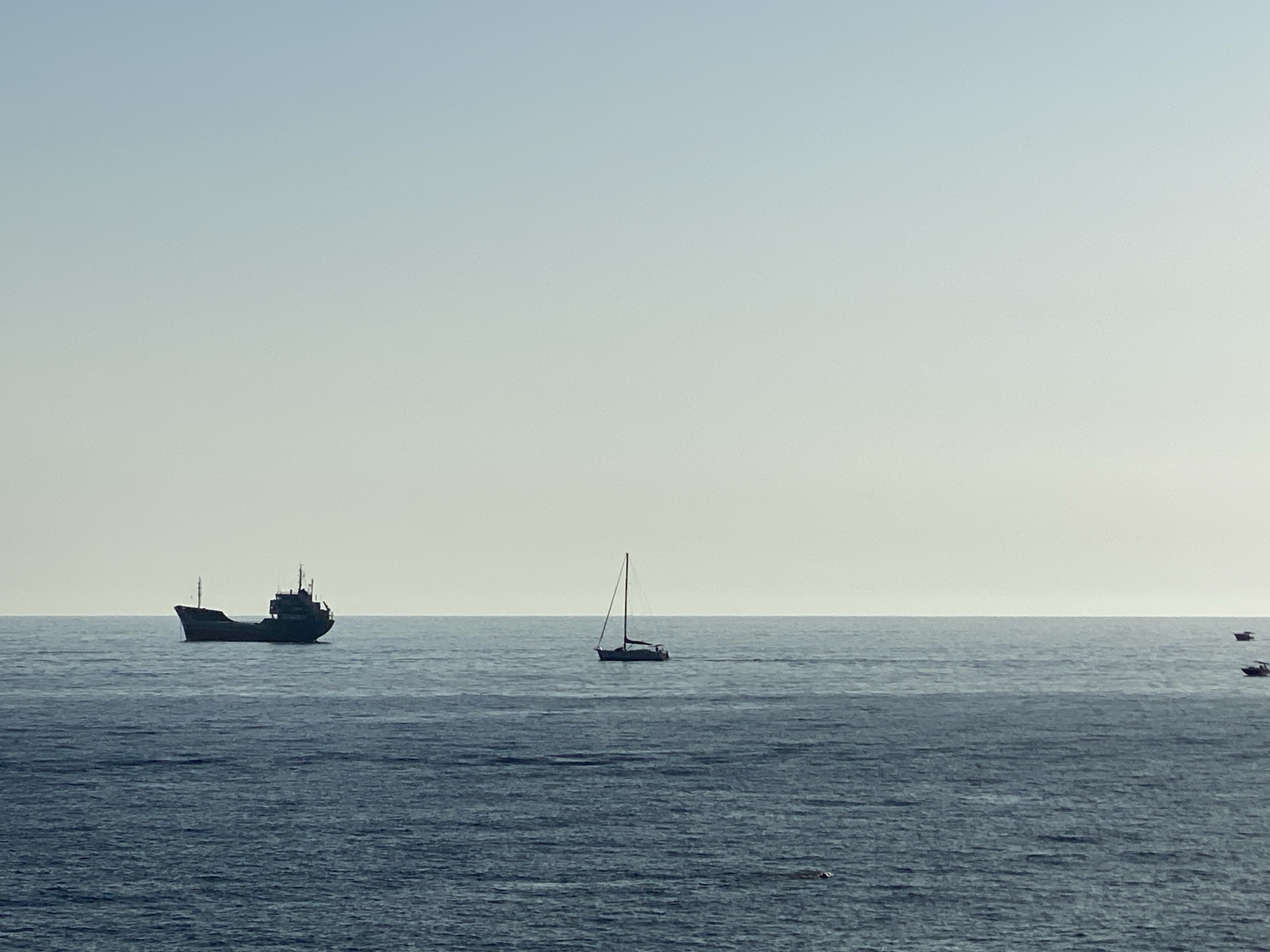 The image depicts a serene seascape with a clear, pale blue sky fading into a lighter hue near the horizon. The sea is a deep blue, with gentle ripples indicating calm conditions. Silhouetted against the horizon, several vessels are visible.
On the left side of the frame, a large cargo ship is anchored. It appears dark and somewhat imposing, with visible superstructure and masts. Further to the right, a solitary sailboat with its mast and sail clearly defined is gliding across the water. In the far distance, to the right, two smaller boats are also visible, adding to the sense of maritime activity.
The scene is likely captured from a vantage point overlooking the Adriatic Sea near Sutomore, Montenegro, a popular coastal town known for its beaches and views of the sea. The overall impression is one of tranquility and open space, characteristic of a peaceful day at sea. There are no people clearly visible in the image, and the focus is entirely on the natural environment and the distant vessels.