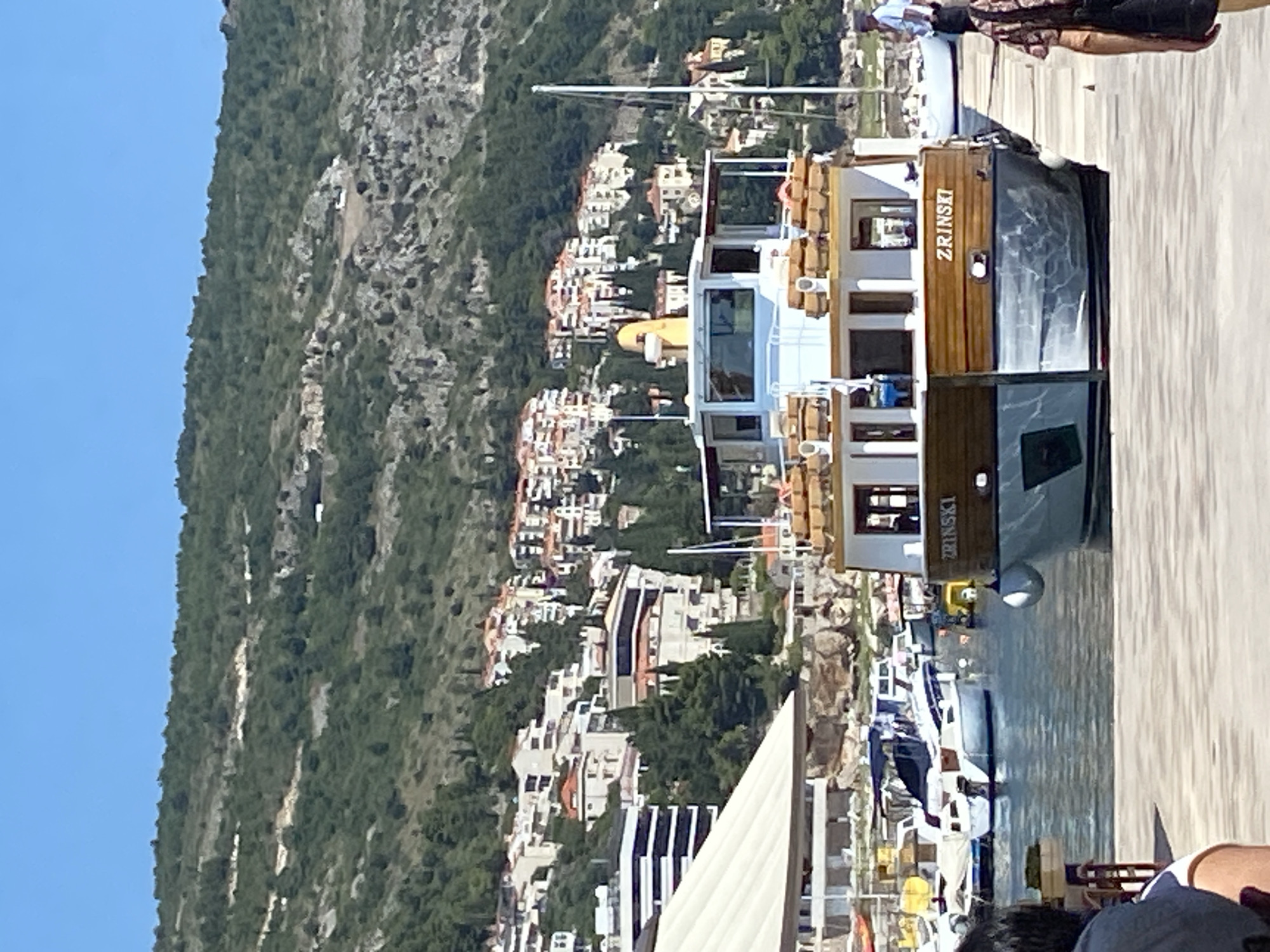 A waterfront scene in Dubrovnik, Croatia, features a cruise boat docked at a pier. The boat, named "ZRINSKI", is white with wooden accents and has multiple decks. In the background, a hillside is densely populated with buildings, many featuring red-tiled roofs, characteristic of Dubrovnik's Old Town architecture. Lush green vegetation covers the upper slopes of the hill. Other boats are visible in the harbor, along with a person standing on the pier and another walking away from the camera. The sky is clear and blue.