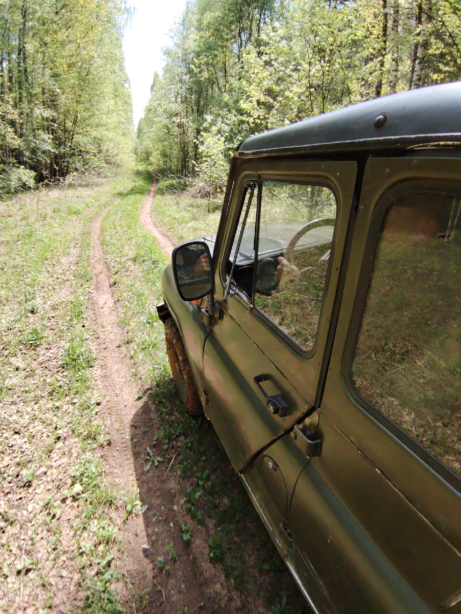 A military-style, green vehicle is parked on a dirt road in a wooded area, possibly in Markovskii, Russia. The road curves into the distance, framed by trees on either side. The vehicle's driver-side window reflects the forest. The environment consists of a dirt road and wooded area. The image suggests a rural or off-road setting.