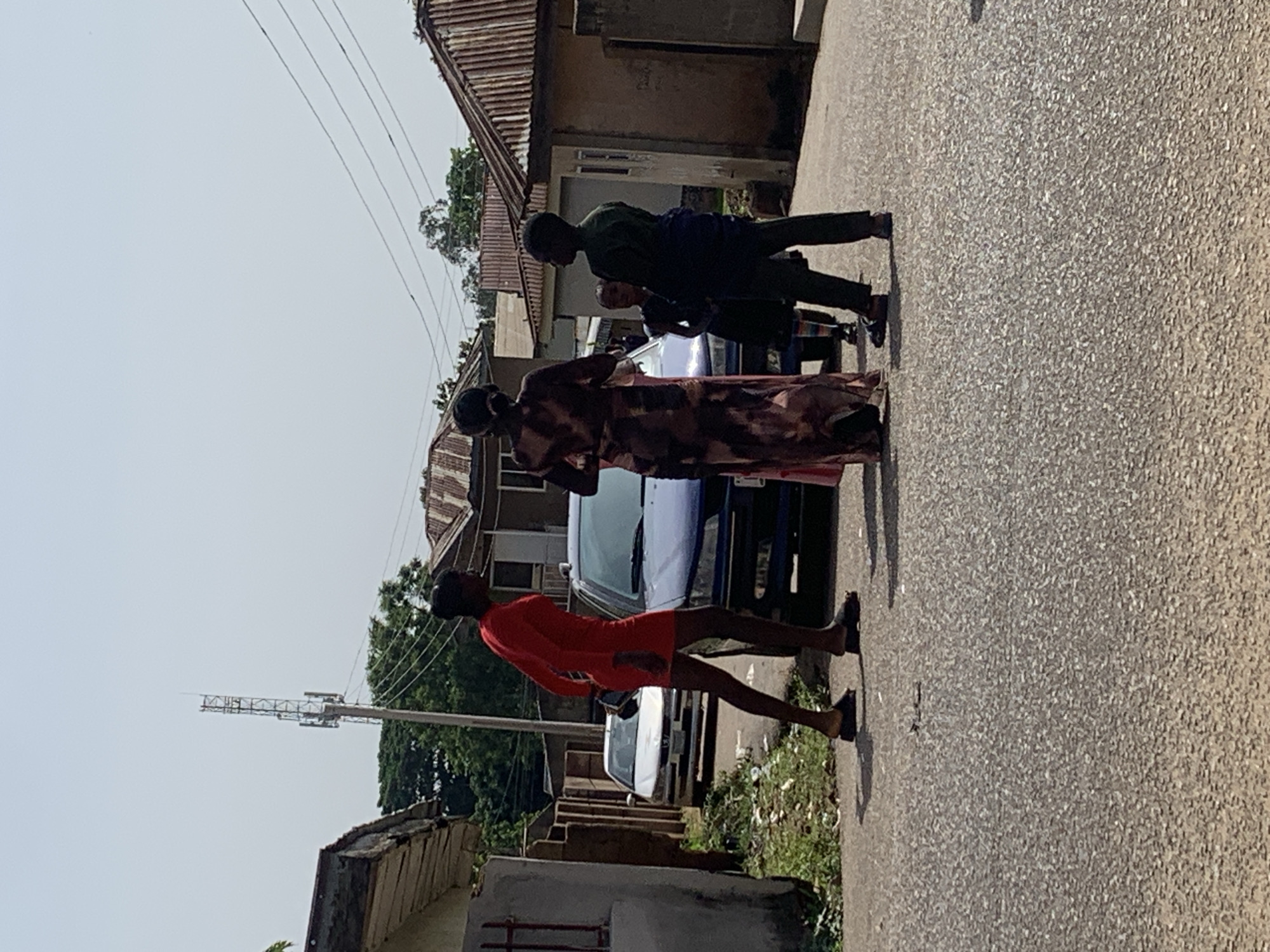 In Gura Top, Nigeria, on a paved street, a group of five individuals are gathered near two parked cars. A woman in a bright red dress stands on the left, facing away from the camera. To her right, a woman in a long, patterned dress is also facing away, engaged in conversation with the others. Three children are present: two appear to be younger, and one older boy in a dark green shirt and black pants stands on the right. The scene is set against a backdrop of residential buildings with corrugated metal roofs, suggesting a typical urban environment. A telecommunications tower is visible in the distance, indicating infrastructure development in the area. The lighting suggests it is daytime, likely mid-morning or afternoon.