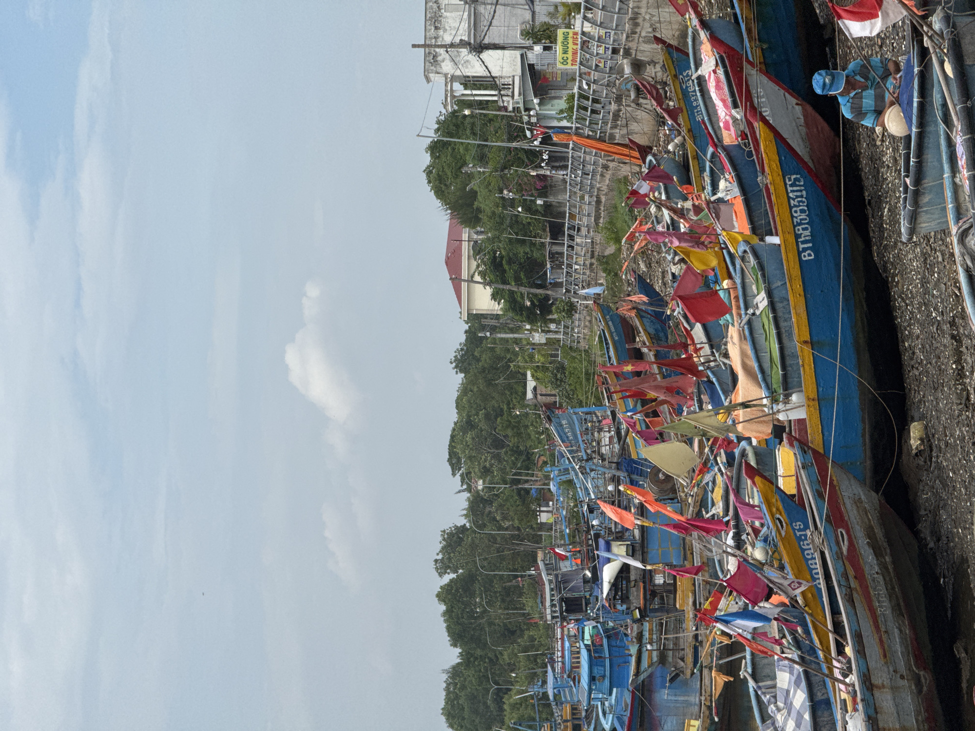 This image depicts a harbor scene in Phú Thủy, Vietnam, crowded with numerous fishing boats. The boats, predominantly blue and yellow, are tightly packed along a gravelly shore. Many are adorned with colorful flags, primarily red and yellow, adding vibrancy to the scene. In the foreground, a person wearing a blue cap and a striped shirt is seated in one of the boats, engaged in an activity with fishing gear. In the background, buildings and lush green trees line the shore, with power lines and streetlights visible. A yellow sign with Vietnamese text is mounted on a wall in the distance. The sky is partly cloudy, with scattered white clouds against a pale blue backdrop. The overall impression is one of a busy fishing community preparing for or returning from sea.