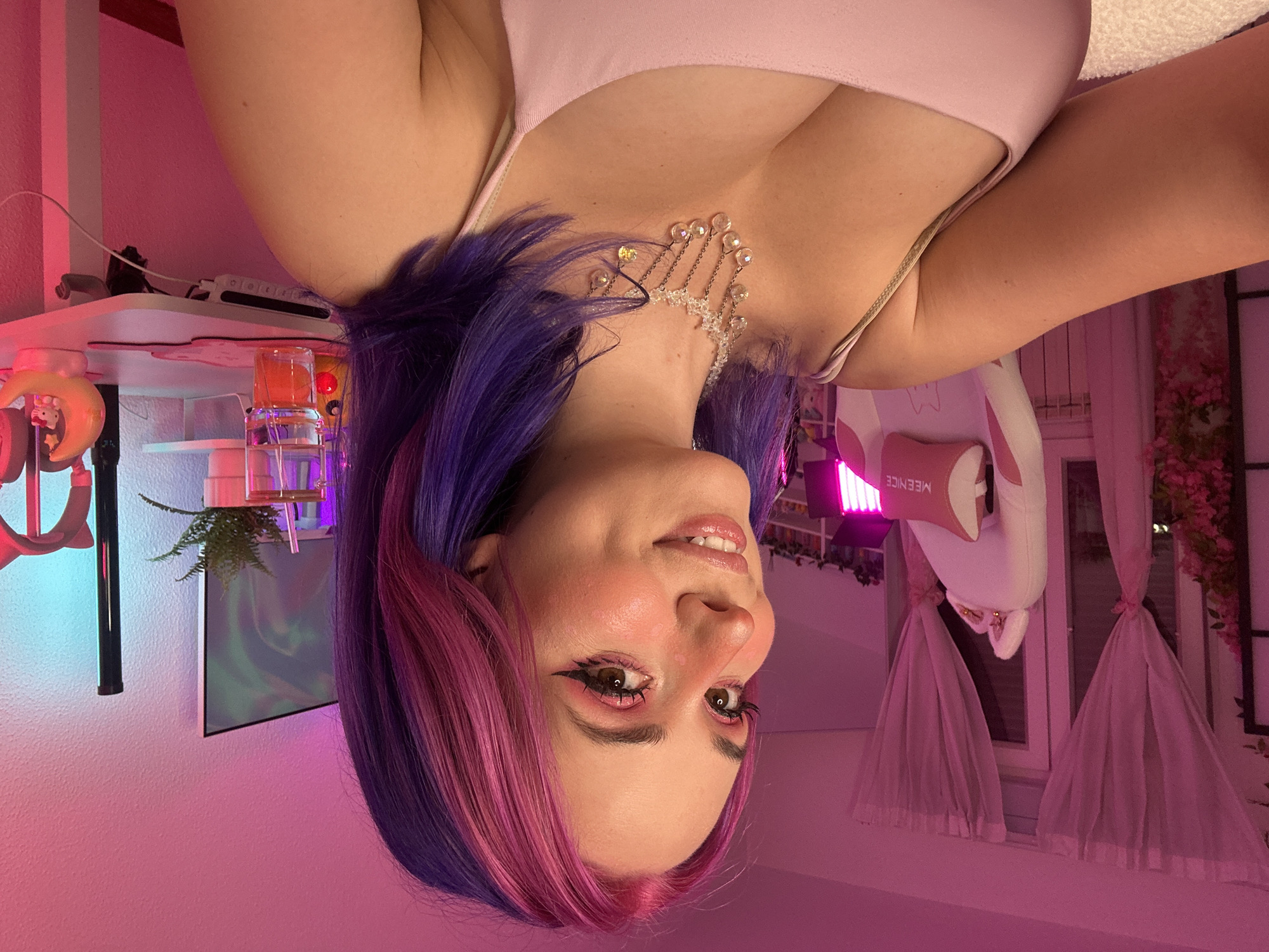 This is a medium shot, taken from the shoulder up, of a young woman with pink and purple dyed hair. She is looking slightly off-camera to her left and smiling. She is wearing a light pink, spaghetti-strap top and a delicate crystal necklace. The lighting is predominantly pink and purple, creating a soft glow in the room.
In the background, there is a pink gaming chair with what appears to be cat ears on the headrest. To the right, a white desk is visible with various items including a plant in a white pot, a glass with a straw and what appears to be an orange slice inside, a monitor displaying a colorful abstract image, and a small crescent moon-shaped night light with Hello Kitty. There are sheer pink curtains hanging behind the chair and a radiator underneath.
The scene appears to be a home office or gaming setup, likely in a residential area. There is no identifiable information suggesting this location is in the city of Oviedo, Spain.
