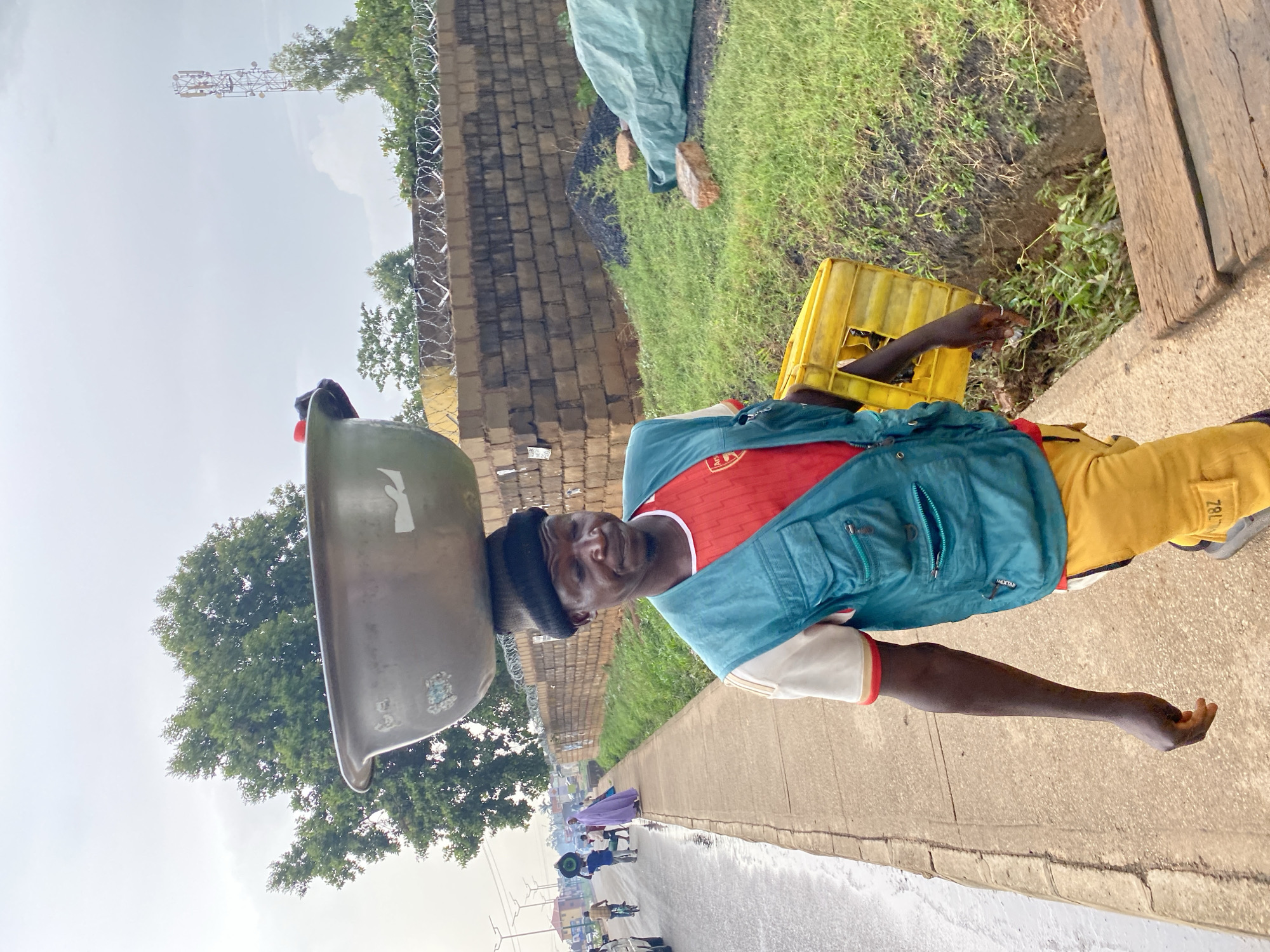 A man in Bukuru, Nigeria, carries a large metal bowl on his head while walking on a sidewalk next to a street. He wears a red t-shirt, yellow pants, and a blue jacket. A yellow crate sits on the ground next to him. In the background, other people walk along the street, and a building with barbed wire on top is visible. A cell tower rises in the distance.