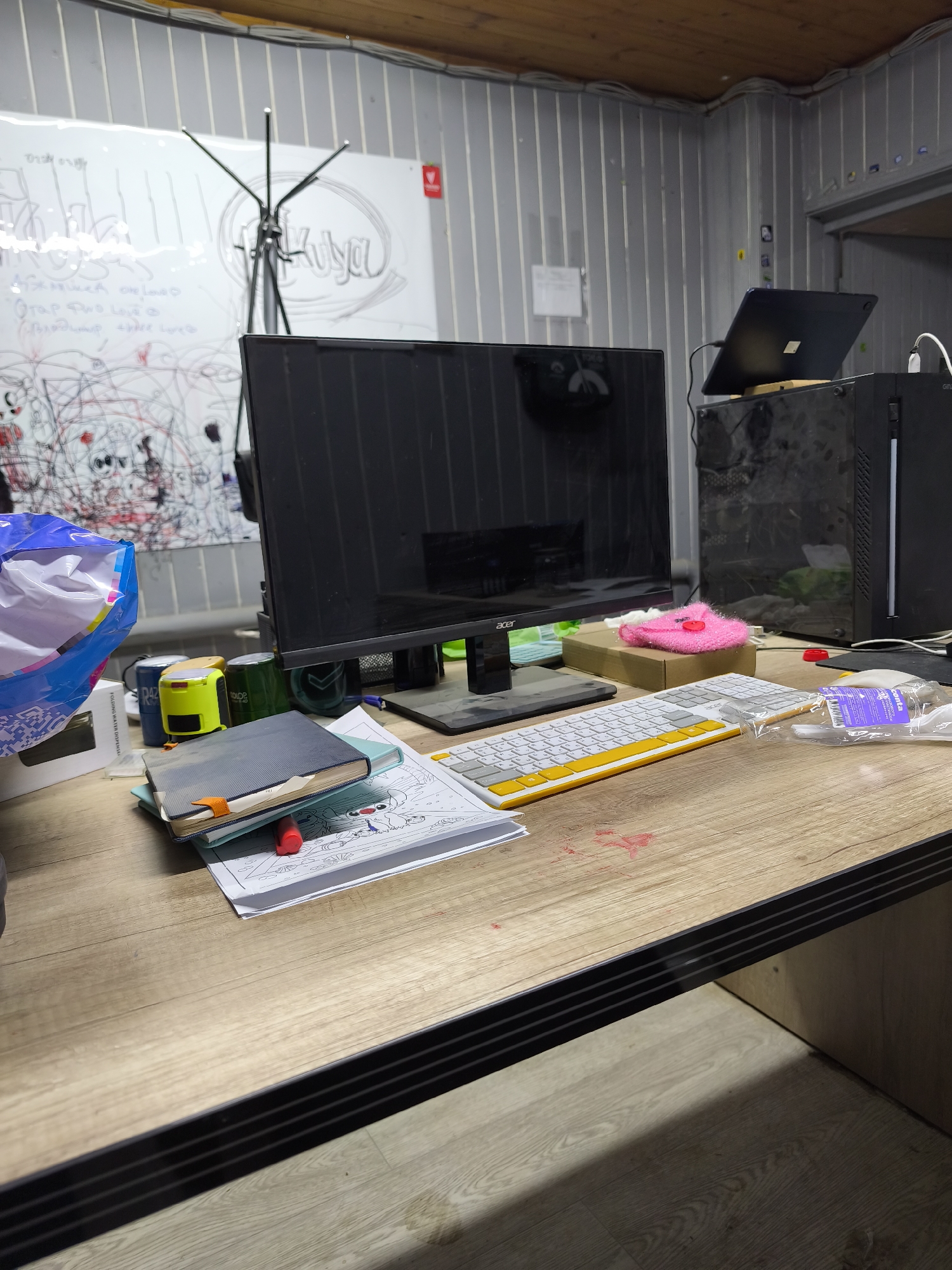 The image shows a cluttered desk in what appears to be an office or workspace. On the desk, there is a monitor, keyboard, laptop, computer tower, notebooks, staplers, a pink fuzzy object, and packaged utensils. The desk has a light wood grain surface with a dark edge. In the background, there is a whiteboard with writing and doodles on it. The walls are metal and corrugated. Based on these details, it is not possible to determine if this location is in Moscow, Russia. The description is limited to the visible contents and characteristics of the scene.