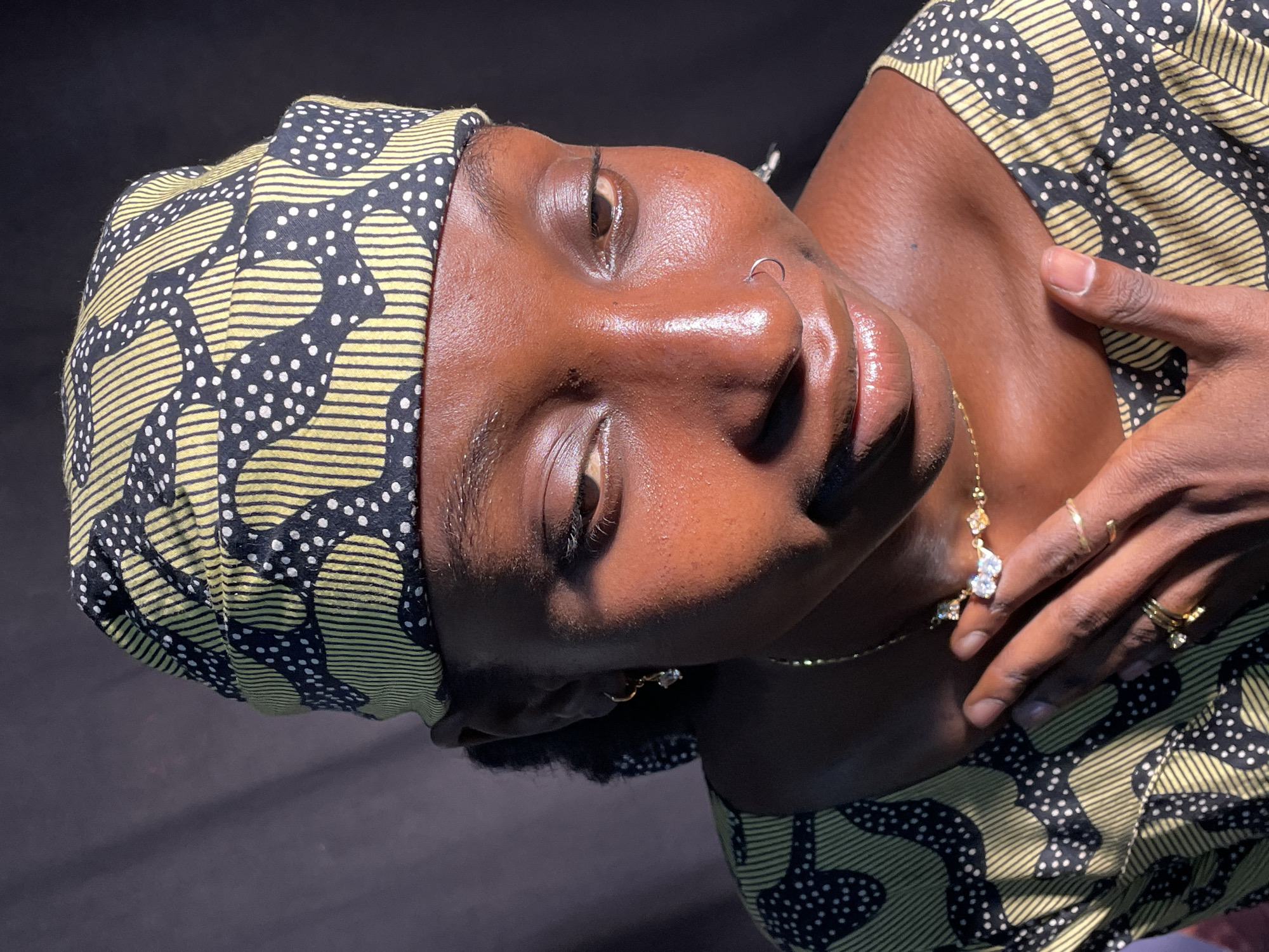 A close-up portrait, taken in Jos, Nigeria, features a person with dark skin, positioned with their head tilted to the left. The subject wears a headwrap and top made of the same fabric, characterized by a striking pattern of black, yellow striped segments, and white polka dots.
Strong, direct lighting, likely sunlight, creates bright highlights on the person's forehead, nose, and lips, accentuating the natural sheen of their skin. Their eyes are gently closed or heavily lidded, conveying a serene and contemplative expression. A minimalist silver hoop nose ring adorns their left nostril, complemented by small, clear-stoned dangle earrings.
Around their neck is a delicate gold chain necklace with ornate pendants, each featuring multiple white stones. The person's left hand, adorned with several gold rings, is gently placed near their collarbone, completing the poised composition.
The background is a uniform dark tone, providing a stark contrast that highlights the subject. The image captures a moment of calm, showcasing the subject's features and intricate accessories. No text is visible within the image.
