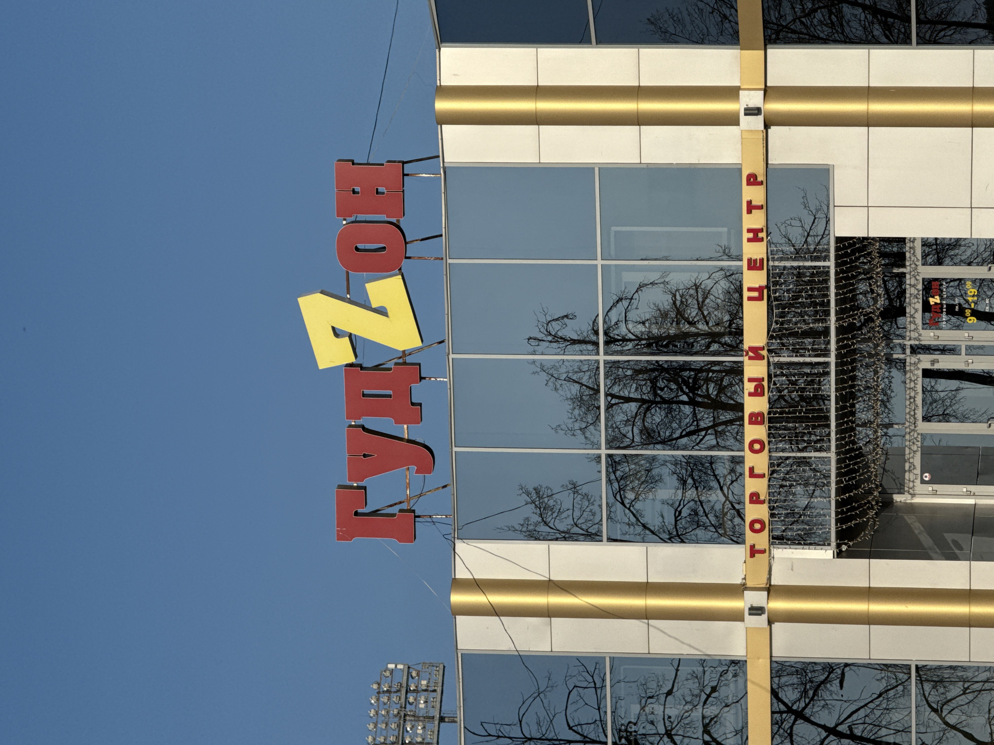 A building with a sign is shown against a blue sky. The sign reads "ГУМЗОН" in red and yellow letters vertically, next to a modern building that appears to be a shopping center. Another sign, oriented vertically, reads "ТОРГОВЫЙ ЦЕНТР" in red letters. Tree branches are reflected in the building's windows. The location is identified as Votkinsk, Russia, based on the Cyrillic text and architectural style. There are utility wires above the building. A light tower is visible on the bottom left corner of the image.