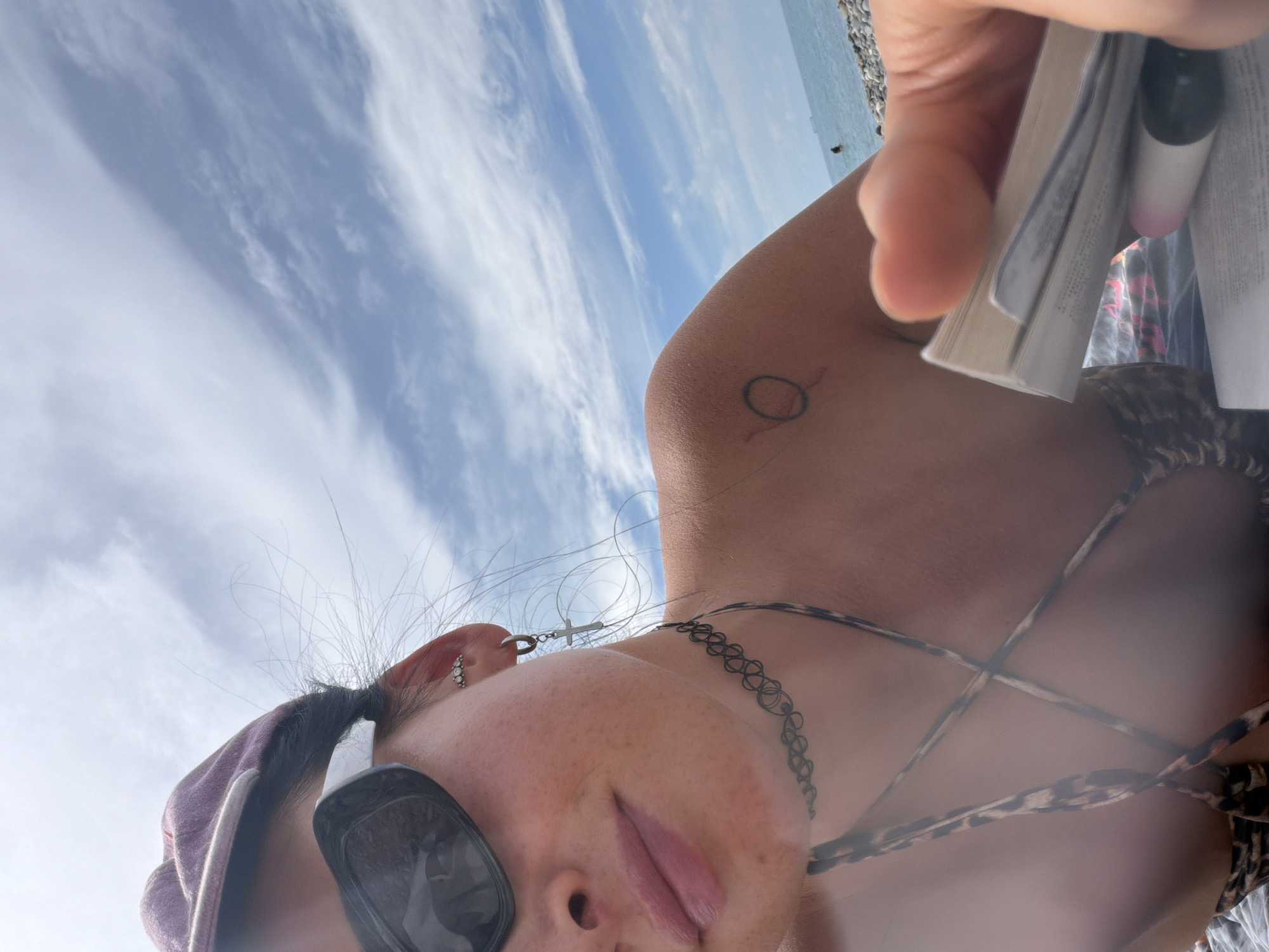 A young woman is lying on a beach in Sochi, Russia, looking up at the sky. She is wearing sunglasses and a bikini top, and has a tattoo on her arm. She is holding a book in her hand. The sky is blue with some clouds, and the sea is visible in the background. The beach is made of pebbles.