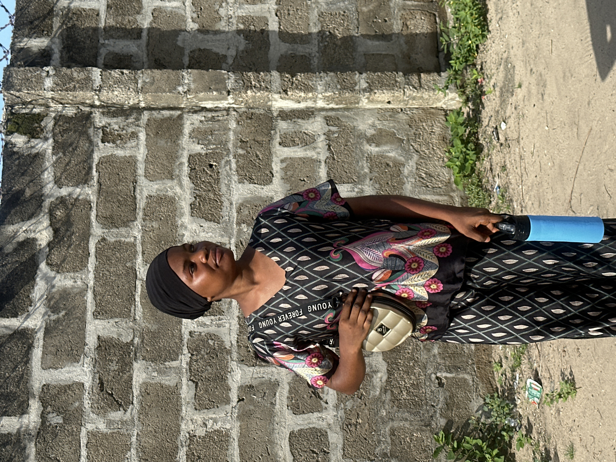 A portrait-oriented outdoor photograph, corrected for its original rotation, depicts a young adult woman standing beside a concrete block wall in Badagry, Nigeria. The woman, who has dark skin and a neutral expression, wears a black head covering and a patterned long dress. The dress features a black background with white diamond and green leaf patterns on the lower half and sleeves, while the upper torso displays a vibrant floral and paisley design in shades of pink, purple, green, and orange. White text across her chest reads "YOUNG FOREVER YOUNG FOREVER."
She holds a light-colored, quilted crossbody bag with a gold clasp in her left hand and a bright blue cylindrical water bottle with a black cap in her right hand. The wall behind her is made of gray concrete blocks with visible mortar, and hints of barbed wire can be seen along its top edge on the left. Green vegetation and weeds grow along the base of the wall, which meets a light brown, dusty dirt path. The strong shadows cast on the wall and ground indicate bright sunlight, suggesting the image was taken during daytime.