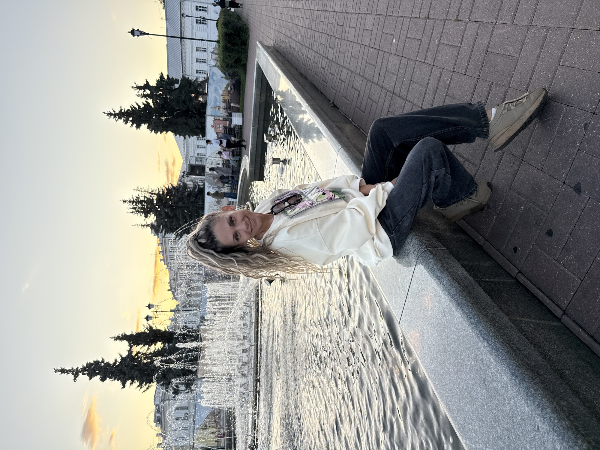 A woman with blonde hair pulled into a high ponytail is sitting on the edge of a fountain in a city square. She is wearing a cream-colored sweatshirt and dark wash jeans, along with tan sneakers. She is smiling at the camera. Behind her, a fountain sprays water, and in the background, there are trees, buildings, and a streetlamp. The sky is a soft yellow and orange, indicating it is either sunrise or sunset. The ground is paved with brick. The location is the city of Rusanovo, Russia.