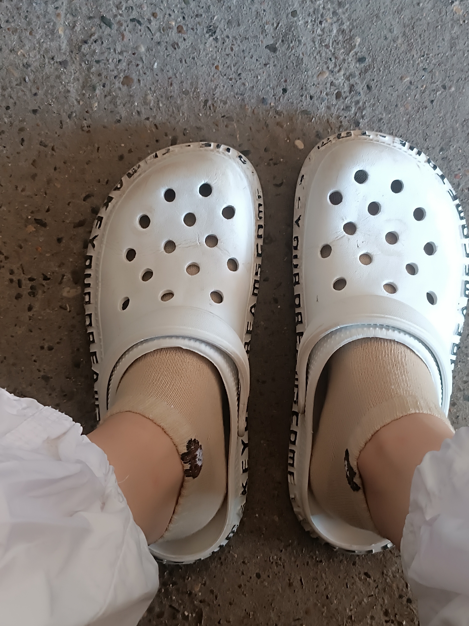 The image shows a pair of white Crocs clogs with tan ankle socks worn on feet. The Crocs have patterned text around the outer edge. The background consists of a gray, textured surface resembling concrete or asphalt. The location appears to be an outdoor setting in an urban area.