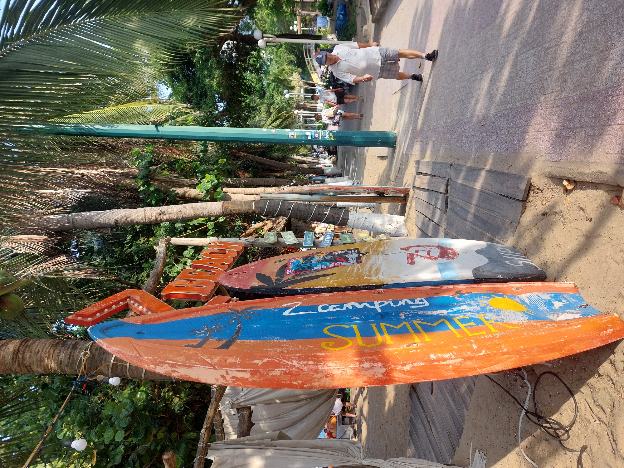 A man in a white polo shirt and plaid shorts walks on a paved pathway lined with palm trees in Nha Trang Ward, Vietnam. He is facing forward and looking slightly to his right. Beside the pathway, on the sand, are several surfboards leaning against a wooden structure with a "BEACH" sign. Curtains hang on the left side of the structure. Other people are visible walking further down the pathway in the distance. The sun is casting shadows from the palm trees onto the pathway.