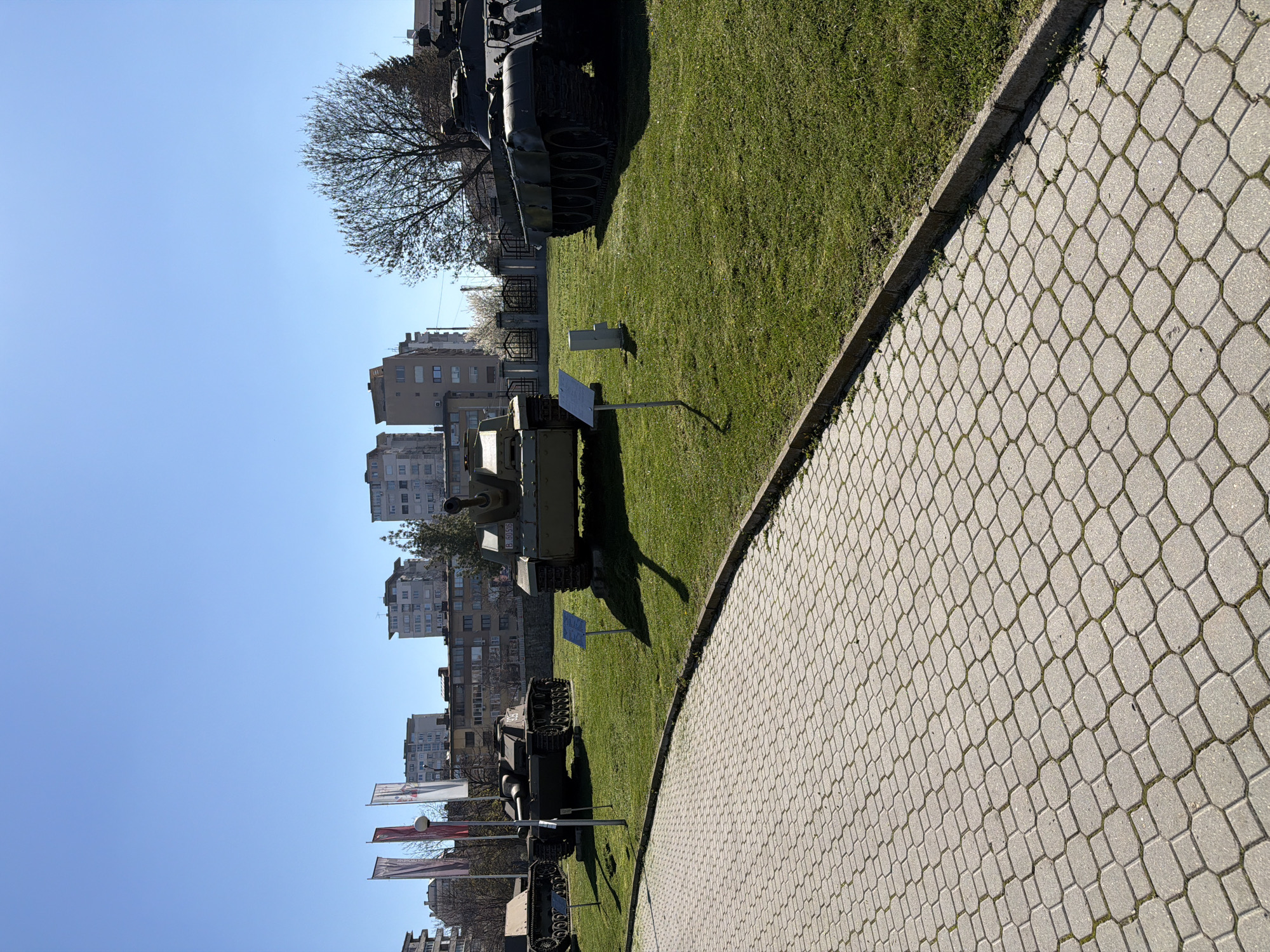 The image shows a military museum in Unknown, Unknown. The primary subjects are several tanks displayed on a grassy area adjacent to a paved walkway with a repeating honeycomb pattern. Buildings stand in the background. Flags are visible near the tanks. A tree with bare branches is to the left of the tanks. The sky is clear and blue.