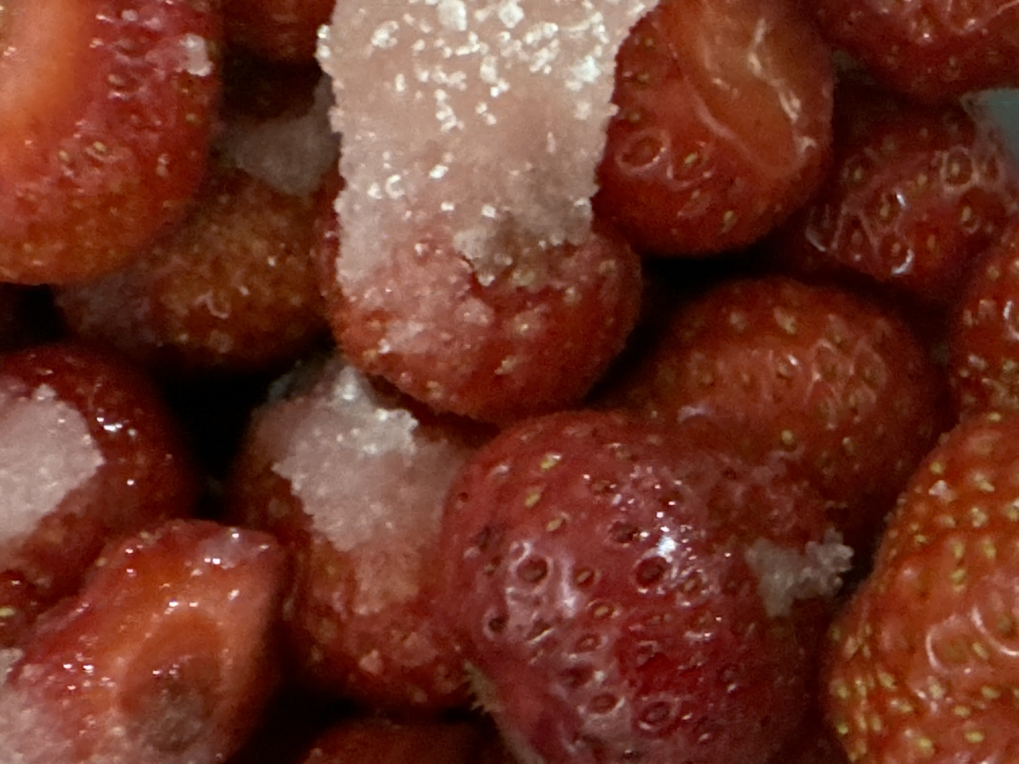 The image shows a close-up, overhead view of a pile of strawberries, some of which are sliced and coated in sugar. The strawberries are a deep red color, and the sugar crystals are white and sparkly. The lighting is soft and diffused, creating a slightly dreamy effect. The background is out of focus, but appears to be a darker, warmer color. There are no people or other objects visible in the image. The location of the strawberries within the city of Waterford, Ireland is not discernible from the image itself.