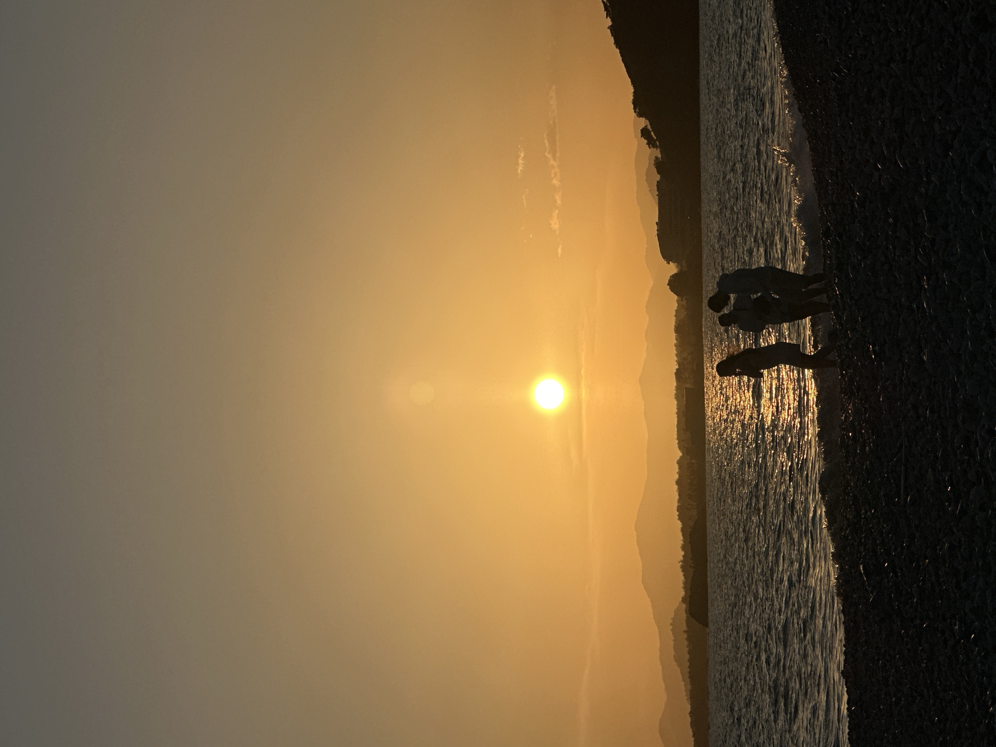 The image shows a sunset over the Mediterranean Sea in Fethiye, Turkey. The sun is low on the horizon, casting a golden light across the sky and water. Silhouetted figures of people are visible on a pebble beach. The coastline is lined with mountains. The interaction between the people and the environment (sea, beach, sunlight) is the primary focus.