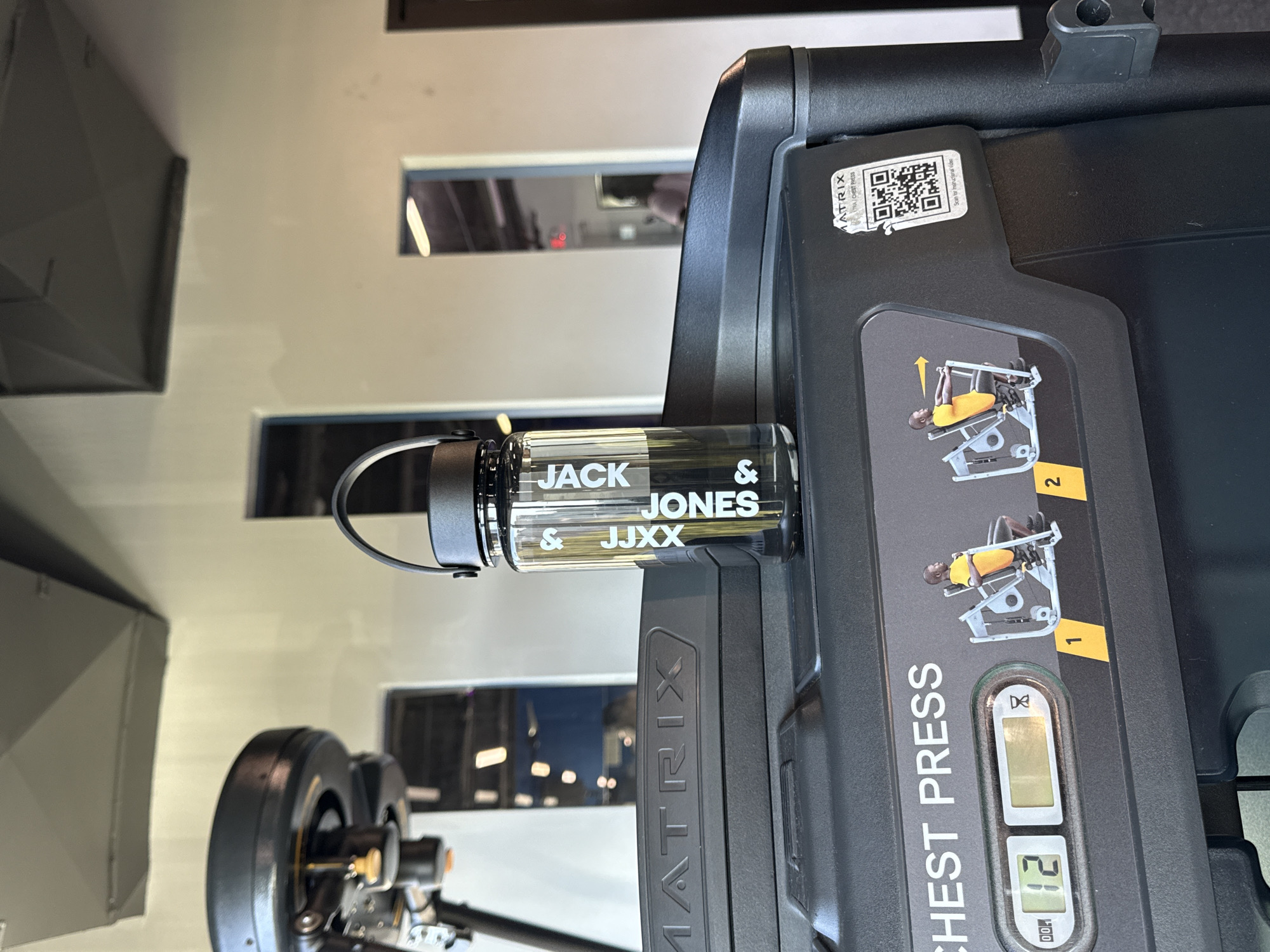 The image is a close-up shot of a black Matrix brand chest press exercise machine in a gym. A clear water bottle with a black lid and handle is positioned on top of the machine. White text on the bottle reads "JACK & JJXX JONES & JJXX". The machine has instructions for use displayed, including illustrations of a person performing the chest press exercise and a digital display showing "12". A QR code with the text "MATRIX ULTRA / CHEST PRESS" and "Scan for Instructional Video" is affixed to the right side of the machine. The background shows a gym environment with white walls, windows, and ceiling structures. The specific location in the City of Yonkers, United States of America, cannot be determined from the image.