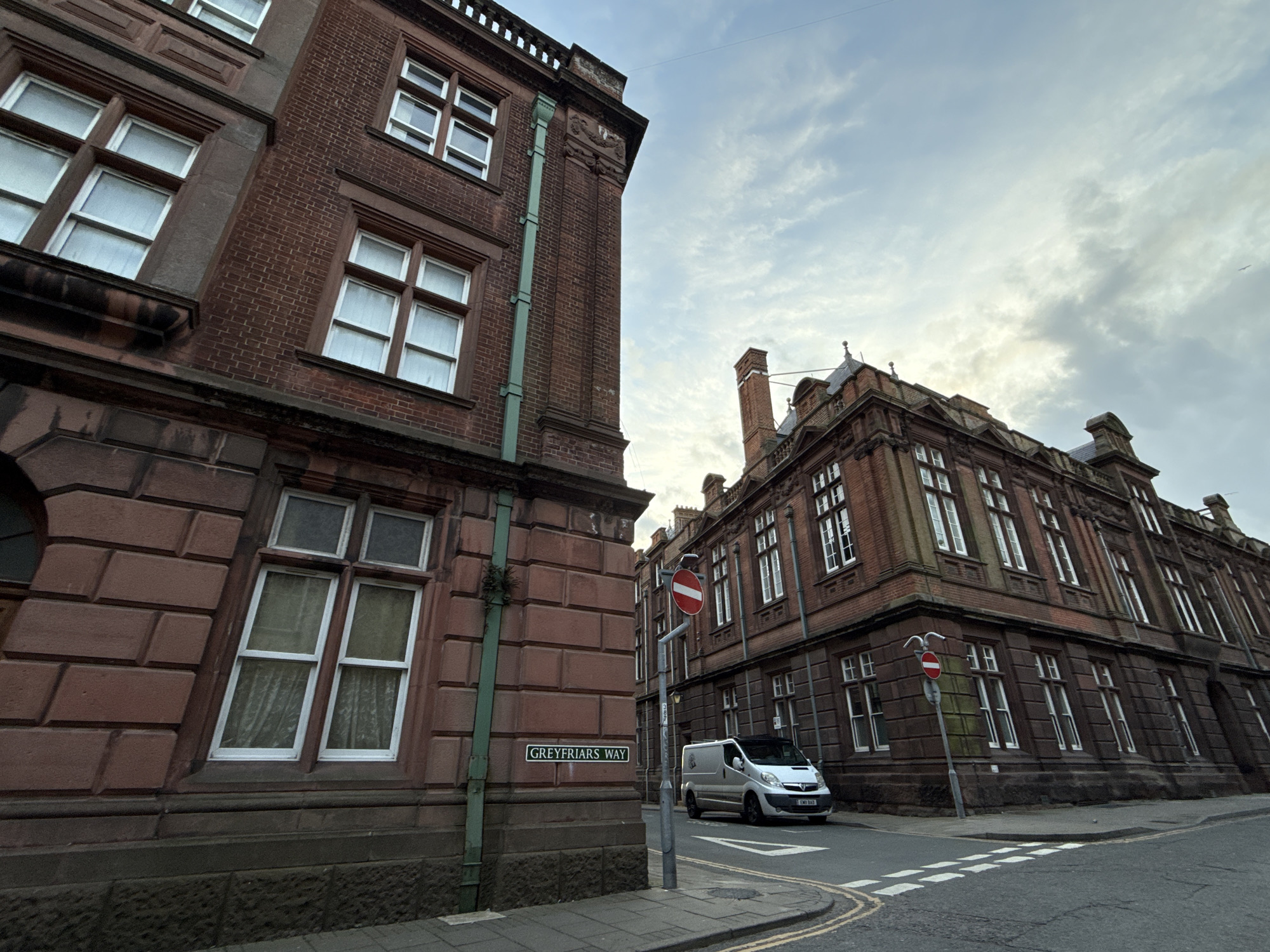 The image presents a street view at the intersection of a street and Greyfriars Way in Great Yarmouth, United Kingdom. The scene is dominated by two large brick buildings with architectural details such as white-framed windows. The building on the left has a green pipe running vertically along its exterior. A street sign reading "GREYFRIARS WAY" is affixed to the corner of this building. Across the intersection, a silver van is parked, and a "no entry" traffic sign is prominently displayed. The street is paved with asphalt and features painted crosswalk lines. The sky is overcast.