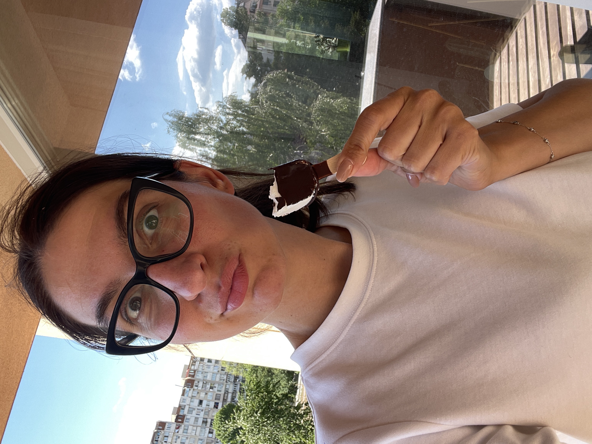 A woman with black-rimmed glasses is holding a partially eaten chocolate-covered ice cream bar in her right hand. She is wearing a white shirt and a delicate bracelet on her right wrist. The background visible through a window shows trees, buildings, and a partly cloudy sky. The architecture of the buildings suggests the image was taken in Sofia, Bulgaria.