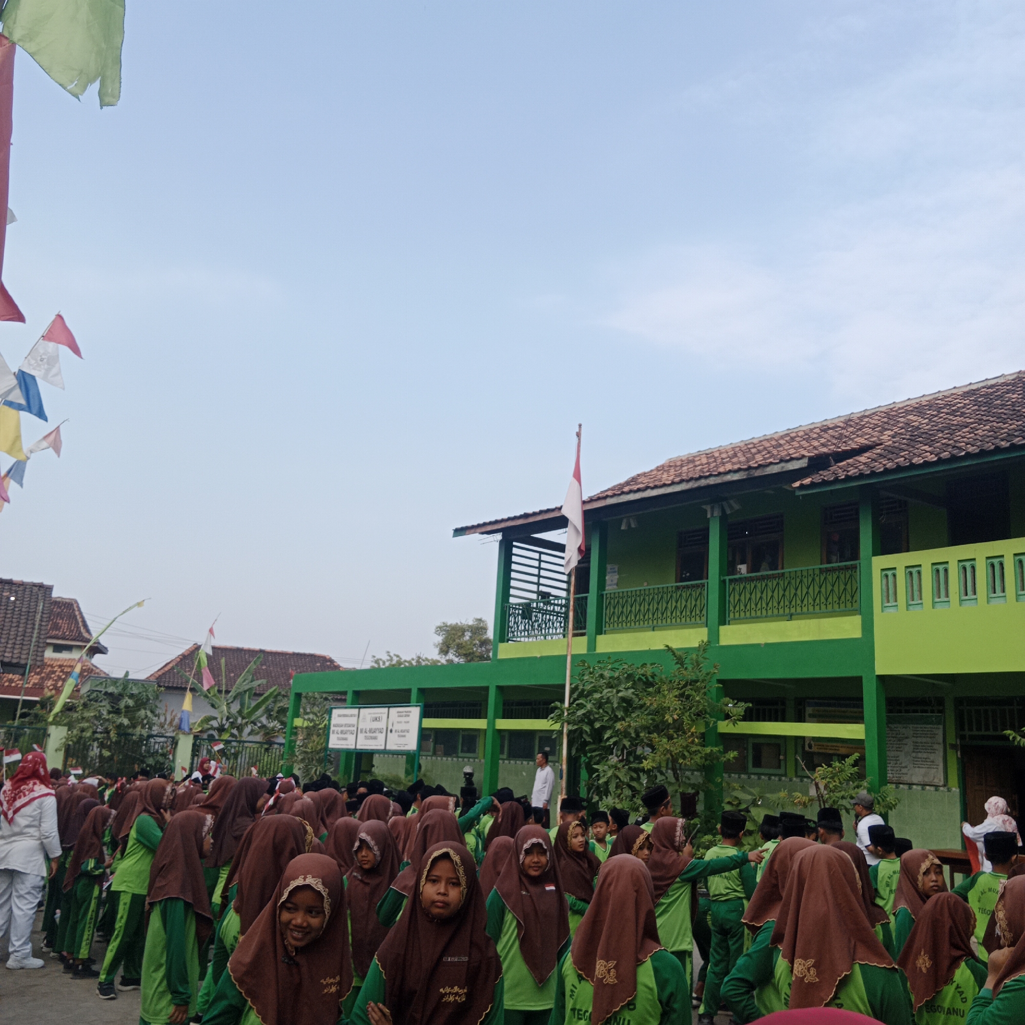 This is a medium shot photo taken outdoors in Tegowanu Kulon, Indonesia. The main subject is a crowd of students, mostly girls, wearing green uniforms and brown hijabs, gathered in front of a two-story green building. The building has a tiled roof, a balcony with decorative railings, and windows. Some students are looking at the camera, while others are looking in different directions.
In the foreground, a girl in a brown hijab and green uniform smiles at the camera. Behind her, more students are visible, some with red and white headscarves. To the left of the frame, a woman in a white uniform and red headscarf stands facing away from the camera.
Flags are strung across the top of the image, including green, red, white, and blue triangular flags. The Indonesian flag, a red and white bicolor, is flying from a flagpole on the building's balcony. The sky is a clear light blue. In the background, other buildings with tiled roofs are visible. A sign with text, possibly a school name, is mounted on the green building. The overall atmosphere suggests an outdoor gathering, likely a school event or assembly.