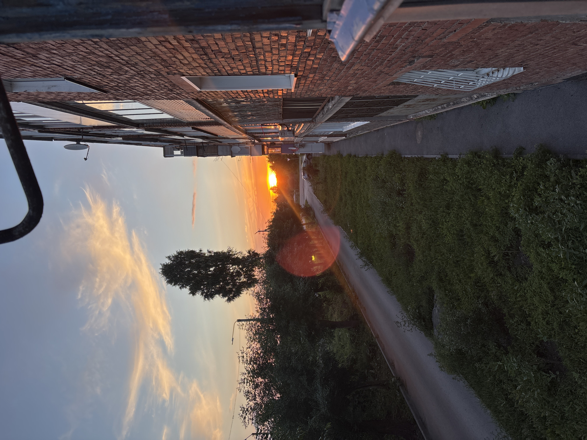 The image depicts an outdoor scene at sunset in Inskoy, Russia. The perspective is from an elevated position, looking down a paved road lined with lush green foliage on the right and a long, brick building with numerous windows on the left. The building appears to be industrial or commercial, with a satellite dish visible on its roof.
The sun is low on the horizon, casting a warm, orange glow across the sky. Wispy, light-colored clouds are scattered across the sky, catching the sunset's light. The road is empty, suggesting a peaceful and quiet evening. A notable lens flare appears in the middle of the image, a circular orange glow caused by the sun. The overall mood of the image is serene and tranquil, capturing the beauty of a sunset over a somewhat industrial landscape. The time of day is clearly evening, and the weather appears clear.