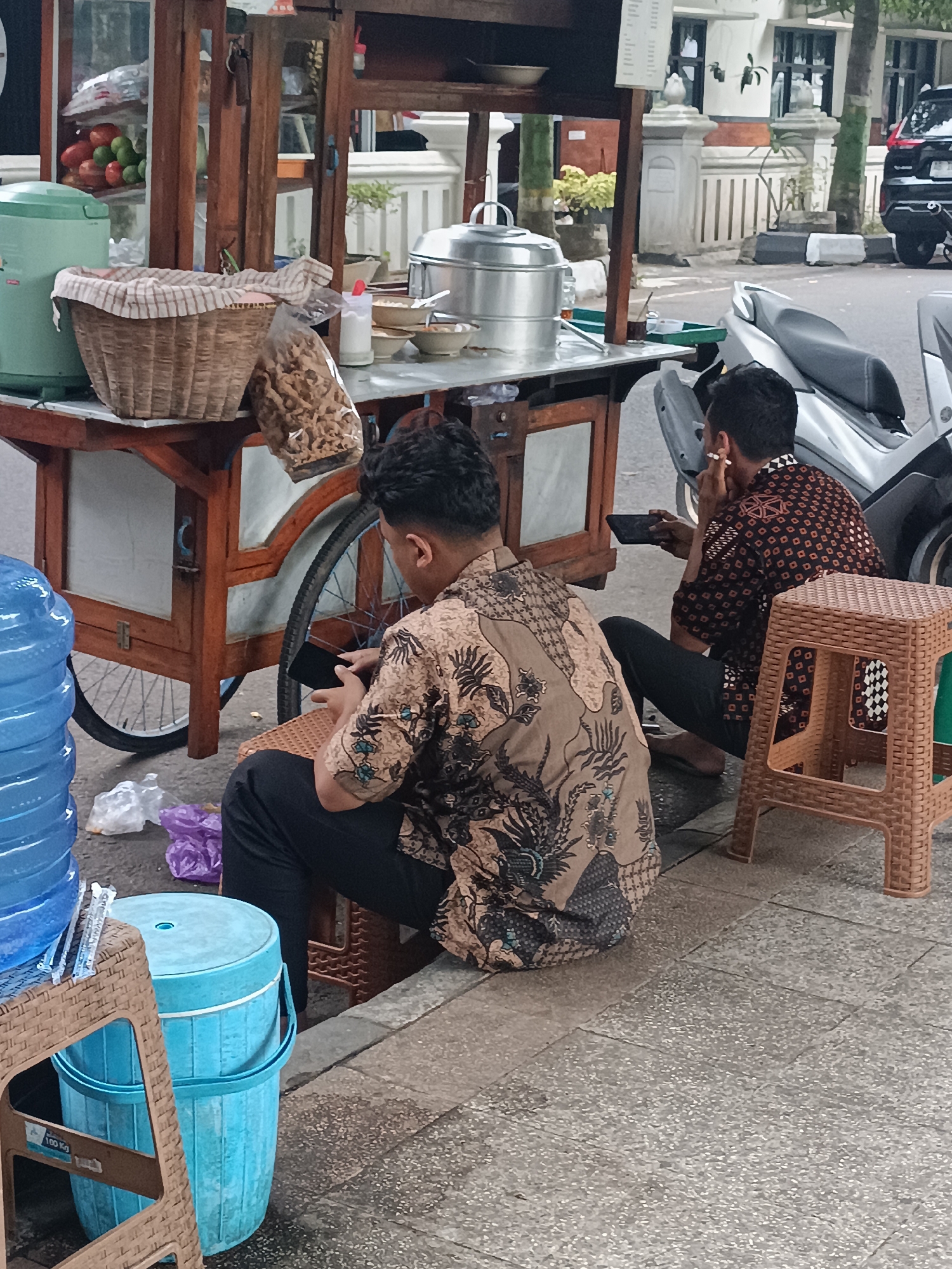 A street food cart is positioned on a sidewalk in Sleman, Indonesia. Two men, dressed in batik shirts, are seated on plastic stools near the cart. The man in the foreground is looking down at a black mobile phone. The man in the background is holding a mobile phone and has a cigarette in his mouth. The food cart contains a basket of snacks, a large green thermos, and what appears to be a steamer pot. Large blue water jugs and a blue cooler are also present on the sidewalk. A motorcycle is parked behind the food cart. The surrounding environment includes a street, trees, and buildings.