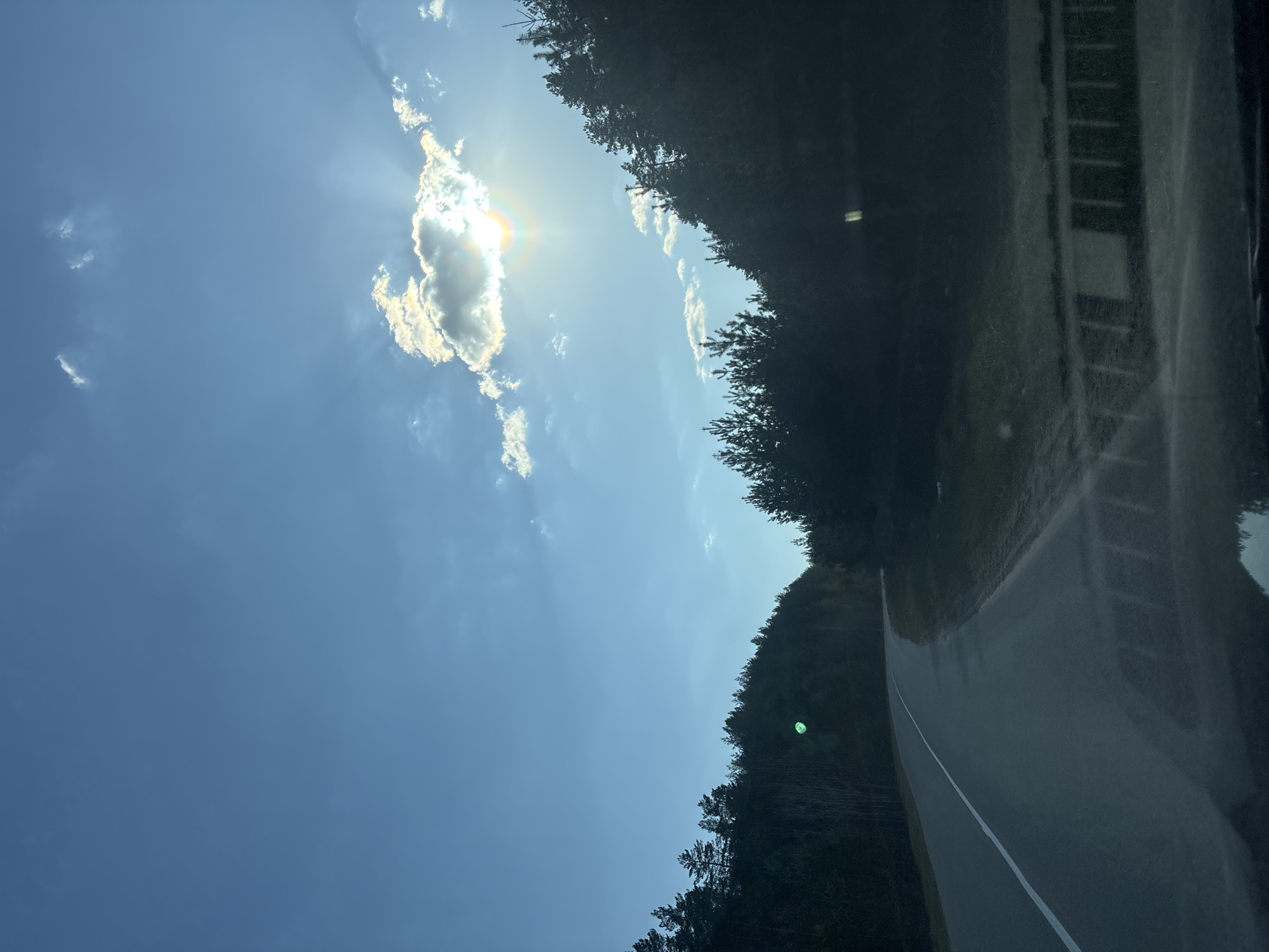 The image shows an asphalt road with a white dividing line running through it. The road is surrounded by dense green trees on both sides. Above, a blue sky is visible with scattered white clouds, and the sun is partially obscured by one of the clouds. It's difficult to determine the exact location in Chaikovsky, Russia, from this image alone. No people are visible.
