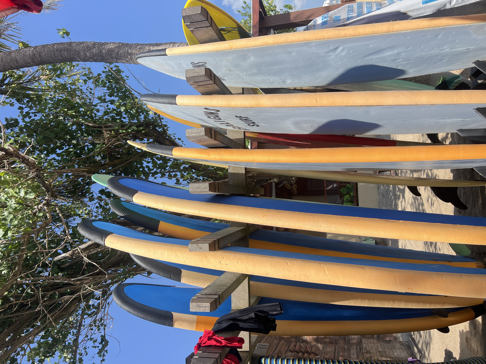 In Kuta, Indonesia, a collection of surfboards stands upright on a sandy surface beneath a dense canopy of trees and a bright blue sky. The surfboards, varying in color from bright blue and yellow to orange and grey, are organized in a wooden rack. Some boards display labels, such as "BALI GUNS SURF." A few items of clothing, a red shirt and a black jacket, are draped over the rack. In the background, a palm tree and a brick wall are visible. The scene appears to be a rental or storage area for surfboards at a beachside location in Kuta.