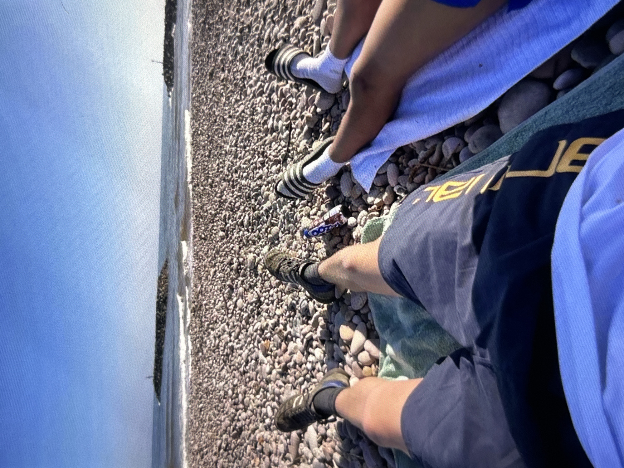 The image shows a high-angle view of two people relaxing on a pebble beach next to the sea under a clear blue sky. The photo is oriented vertically, with the horizon line running down the right side of the frame.
In the foreground, the legs of two individuals are visible. One person, to the right, wears dark blue shorts with yellow lettering that appears to spell "JEAL" (possibly part of a word like "JEALOUS" or a brand name) on one leg. They are wearing dark athletic shoes and dark socks and are sitting on a green towel. The second person, slightly behind and to the left, wears light-colored shorts, white socks, and black and white striped slides. They are resting on a light blue towel.
A small blue and white package, possibly a snack wrapper, with the word "KOOZA" partially visible, lies on the pebbles near the feet of the person in slides. The beach consists entirely of small, grey and white pebbles that extend towards the calm, blue sea. In the distance, a dark structure, likely a breakwater or jetty, protrudes into the water. The sky is bright and cloudless, indicating sunny weather during the day. The scene depicts a leisurely moment on a coastal pebble beach.
