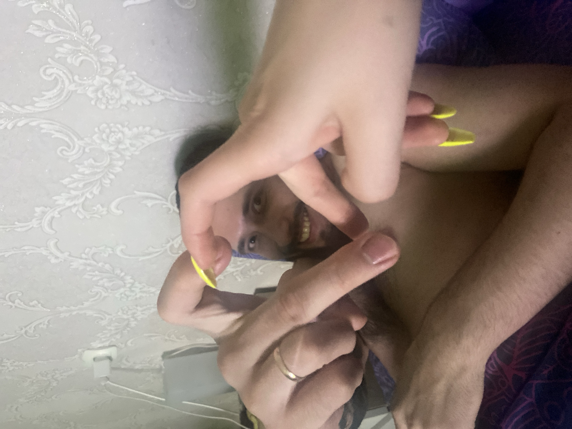 A man with dark hair and a beard smiles into the camera while lying down. Two hands are in the foreground, one holding up two fingers with long yellow nails. The other hand has a gold band on the ring finger. The background is a wall with floral wallpaper and a white electrical outlet. The photo was taken indoors, location unknown, but does not appear to be Chaikovsky, Russia.
