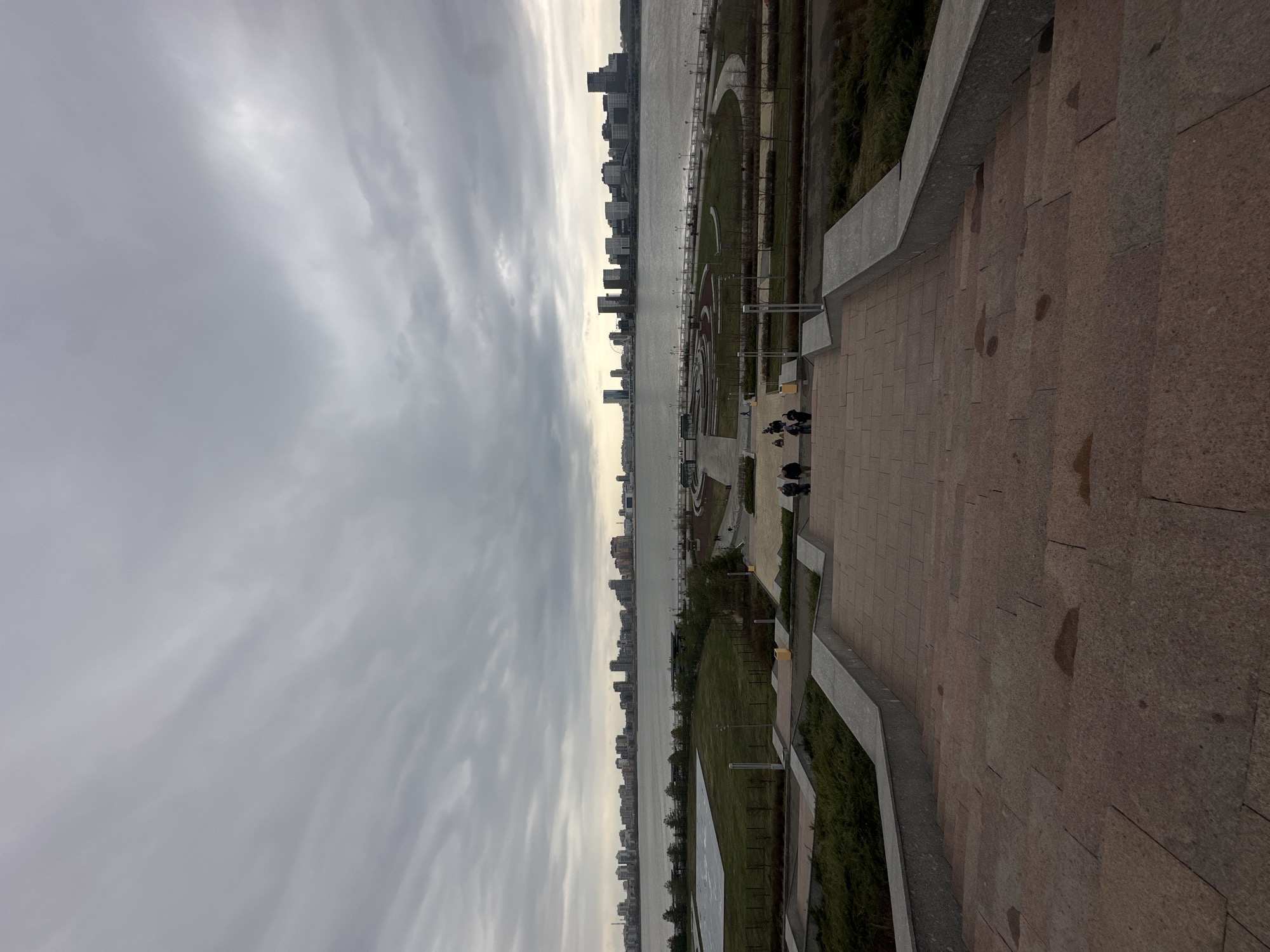 View from a high vantage point in Kazan, Russia, overlooking the waterfront. The camera is positioned on a paved terrace with concrete railings. Below, there is a well-maintained green space with paths, lawns, and young trees. Beyond the green space is the river, and on the opposite bank is a skyline of high-rise buildings under a cloudy sky. A small group of people is gathered in the walkway below.