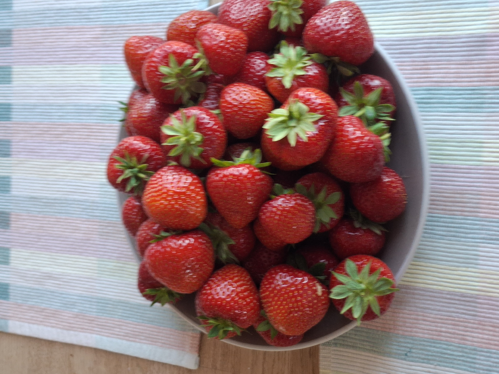 A close-up, high-angle shot shows a grey ceramic bowl filled with fresh, ripe strawberries. The bowl is placed on a striped placemat with pastel pink, yellow, and green stripes. The placemat is on a wooden surface. The strawberries are a vibrant red and appear to have visible seeds and green leafy tops. Some strawberries are whole, while others are cut in half, revealing their texture. The lighting suggests an indoor setting, likely a kitchen or dining area. There are no people present in the image, and no specific location in Åsgårdstrand, Norway can be identified.
