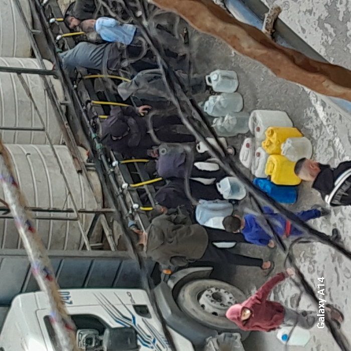 The image displays an outdoor scene in Khan Yunis, Palestinian Territory, focused on a water distribution operation. A white commercial truck, marked "240" on its cab, is positioned on a paved surface. Its trailer section is equipped with multiple large, horizontal cylindrical tanks and a side-mounted manifold system featuring numerous spigots and yellow hoses. Approximately 10-15 individuals, comprising adults and several children, are clustered around this dispensing system. They are actively engaged in filling a variety of plastic containers, including clear, white, blue, and yellow jerrycans and bottles, with water from the spigots. Some individuals are seen holding containers under the spigots, while others are carrying filled containers away or waiting. A person in a maroon hooded top walks with a small container, and another in a blue tracksuit carries a similar item. Numerous full and empty plastic containers are arranged on the ground adjacent to the truck. The scene is observed from an elevated perspective, with dark wires diagonally crossing the foreground.