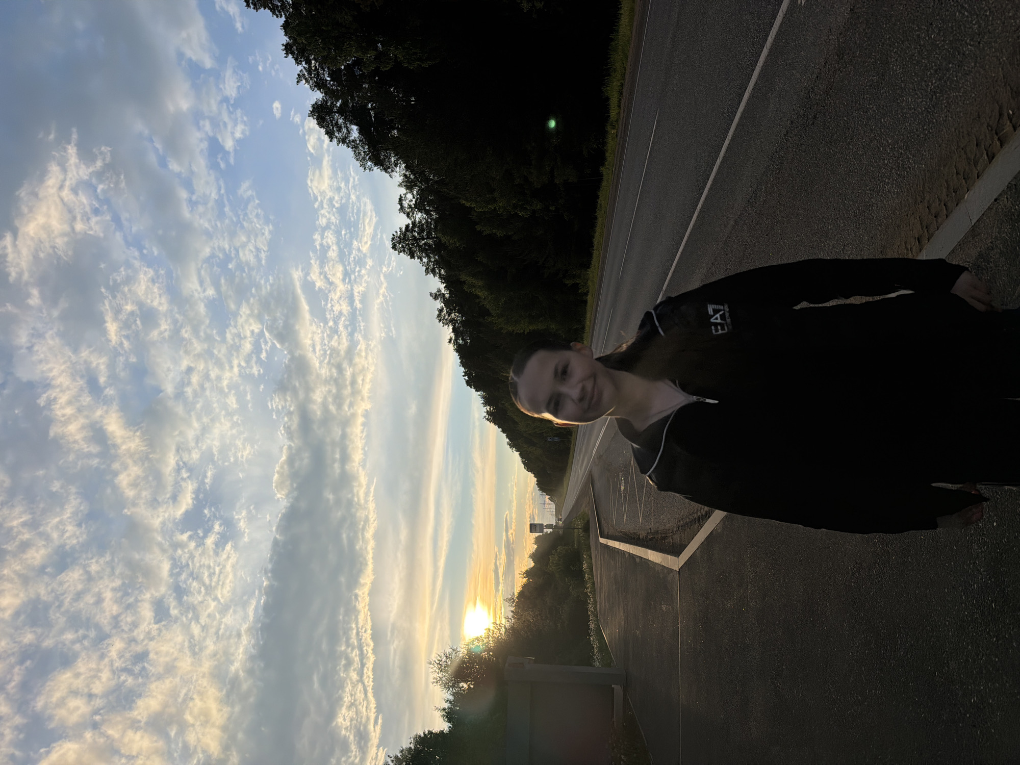 A person stands on the side of a road near Chaikovsky, Russia at sunset. The sky is a mix of blue and white clouds, with the sun setting in the distance, casting a golden glow. On the right side of the frame is a road, with a forest bordering it. The person is wearing a black jacket with "EAT" printed on it.