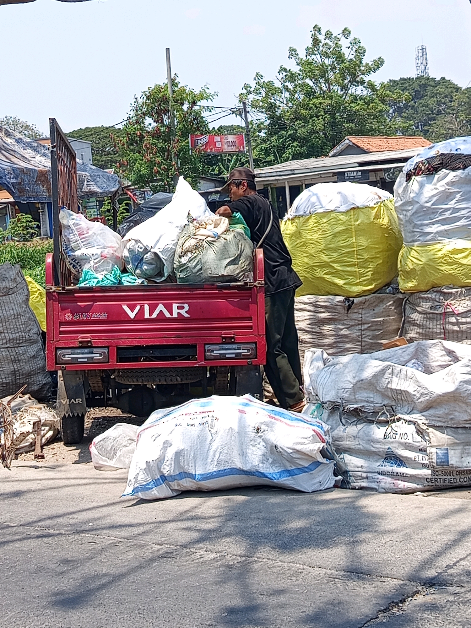 The image depicts a man in a dark shirt and a hat standing on the street next to a red pickup truck. The truck is loaded with various bags, some white, some green, and some clear plastic containing what appears to be recyclables. The man is reaching into the truck bed, interacting with the contents. In the background, there are buildings, trees, and a cell tower, suggesting an urban or semi-urban environment. A prominent sign for "VIAR" is visible on the truck. The ground is paved, and several large sacks are piled up on the right side of the image, some are yellow and white. The overall scene suggests a person engaged in collecting or distributing goods, possibly related to recycling or waste management, in Karawang, Indonesia.