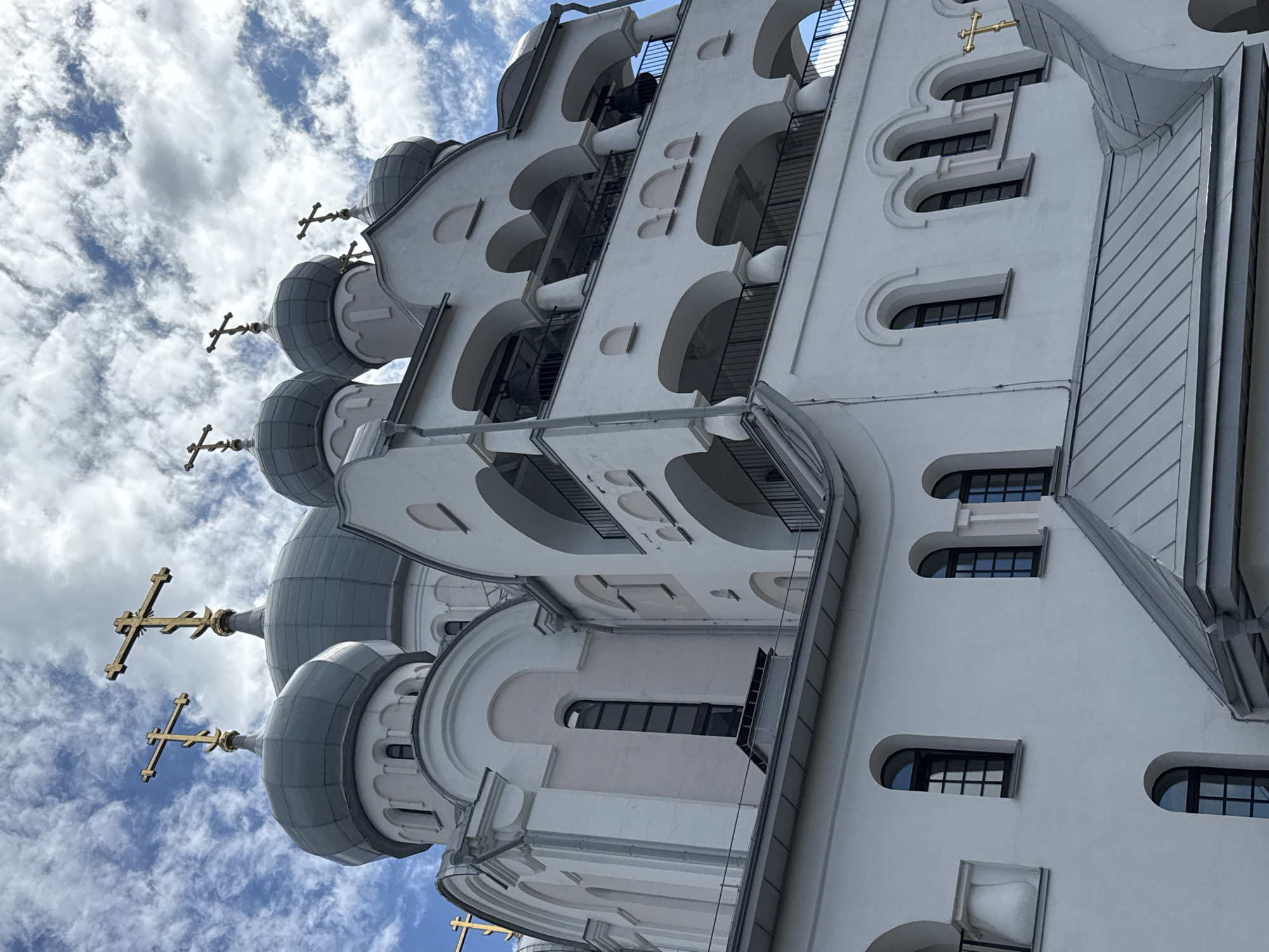 This image shows the exterior of a white Orthodox church with multiple grey domes, each topped with a golden cross. The church architecture features arched windows and decorative elements, including a bell tower with multiple levels of arcades. The sky is a mix of blue with white, fluffy clouds, suggesting it is daytime. There are no people visible in the frame. The church appears to be an active religious building, likely located in Saint Petersburg, Russia, given the architectural style.