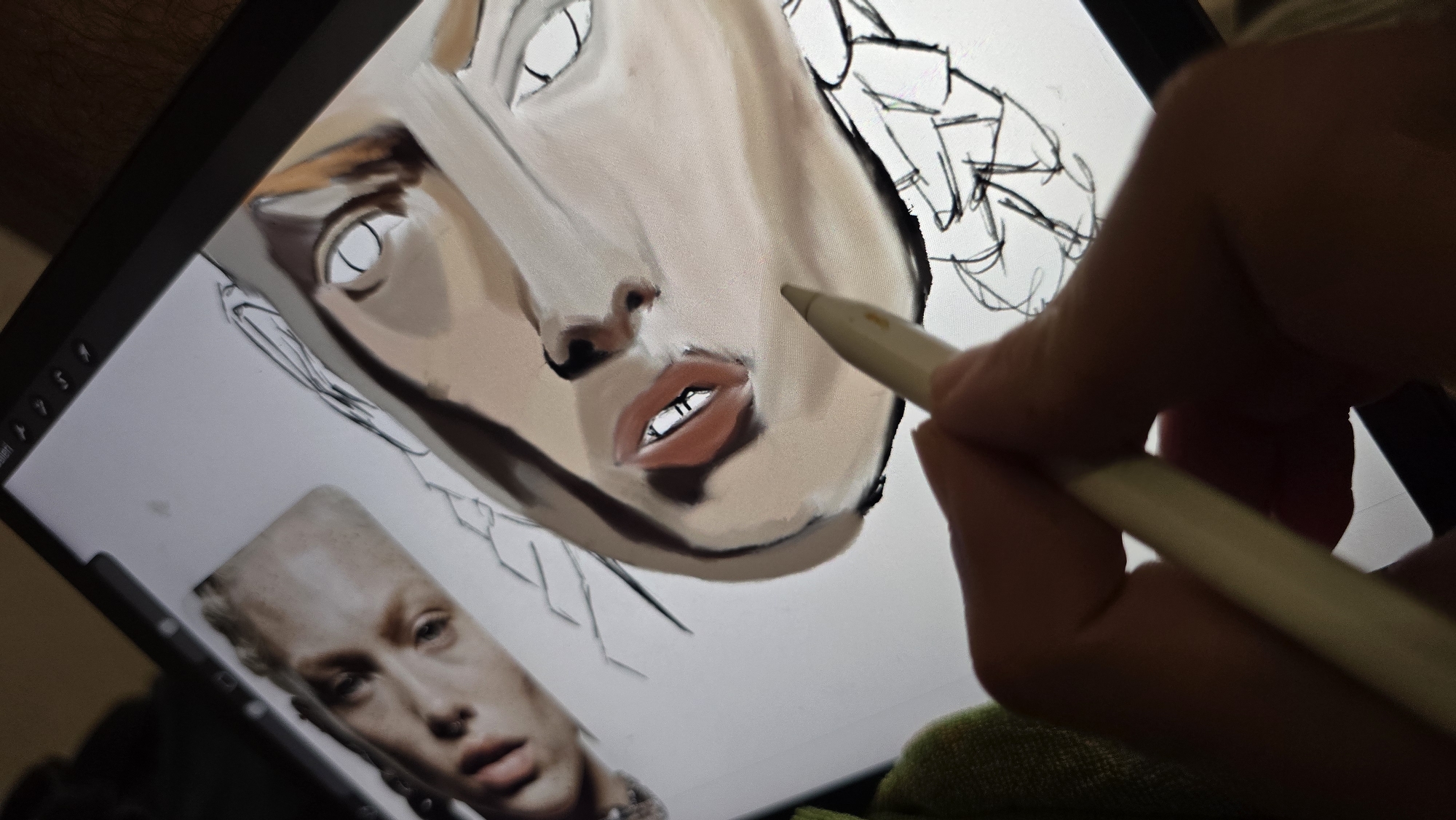 An overhead shot shows a hand holding a stylus, drawing on a tablet. The tablet displays a digital painting of a face. To the left of the painting, a photograph of another face is visible, seemingly a reference image. The person is using a stylus to add shading and detail to the digital artwork. The background is dark and indistinct, with a hint of green fabric on the left side, suggesting the user might be seated. There is no information about the location of this scene in Tolyatti, Russia.